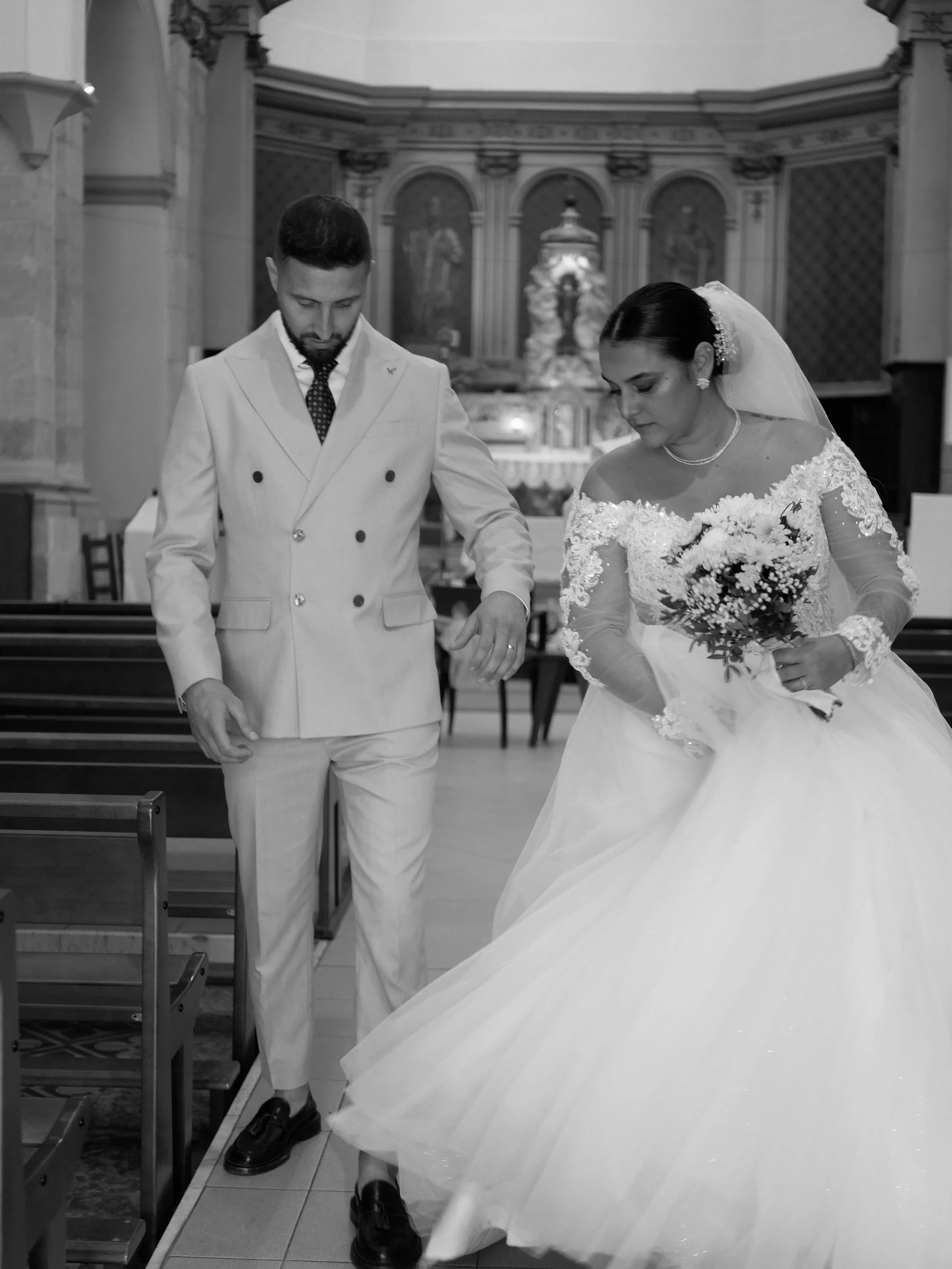 Un couple de mariés dans une église. La mariée porte une robe de mariée avec dentelle et un voile, et tient un bouquet de fleurs. L'homme porte un costume clair avec une cravate à pois.