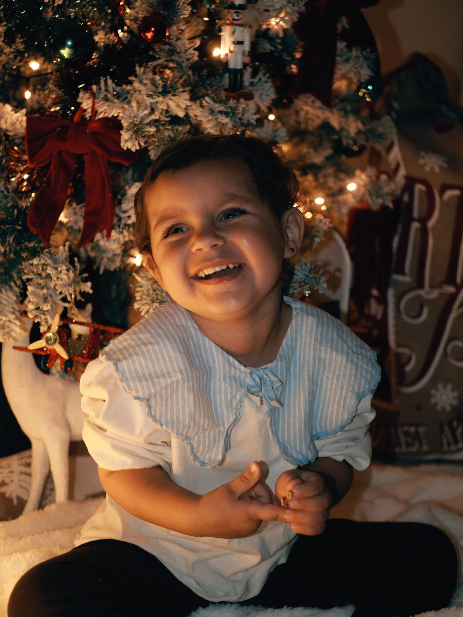 Un enfant souriant assis devant un sapin de Noël décoré avec des boules, des rubans rouges et des lumières, en famille.