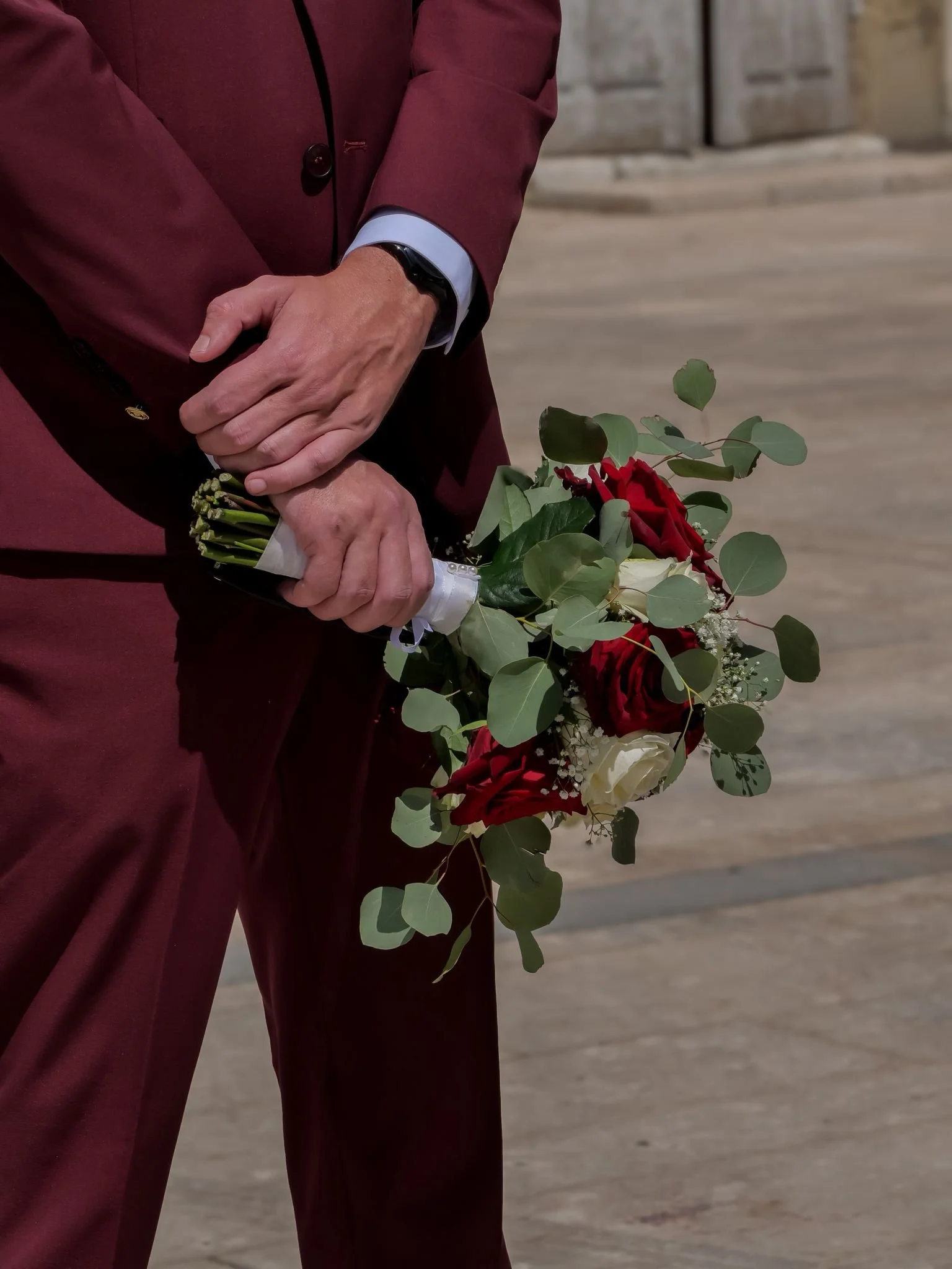 Une personne en costume bordeaux tient un bouquet de fleurs composé de roses rouges et blanches, agrémenté de feuilles vertes, dans un environnement extérieur.