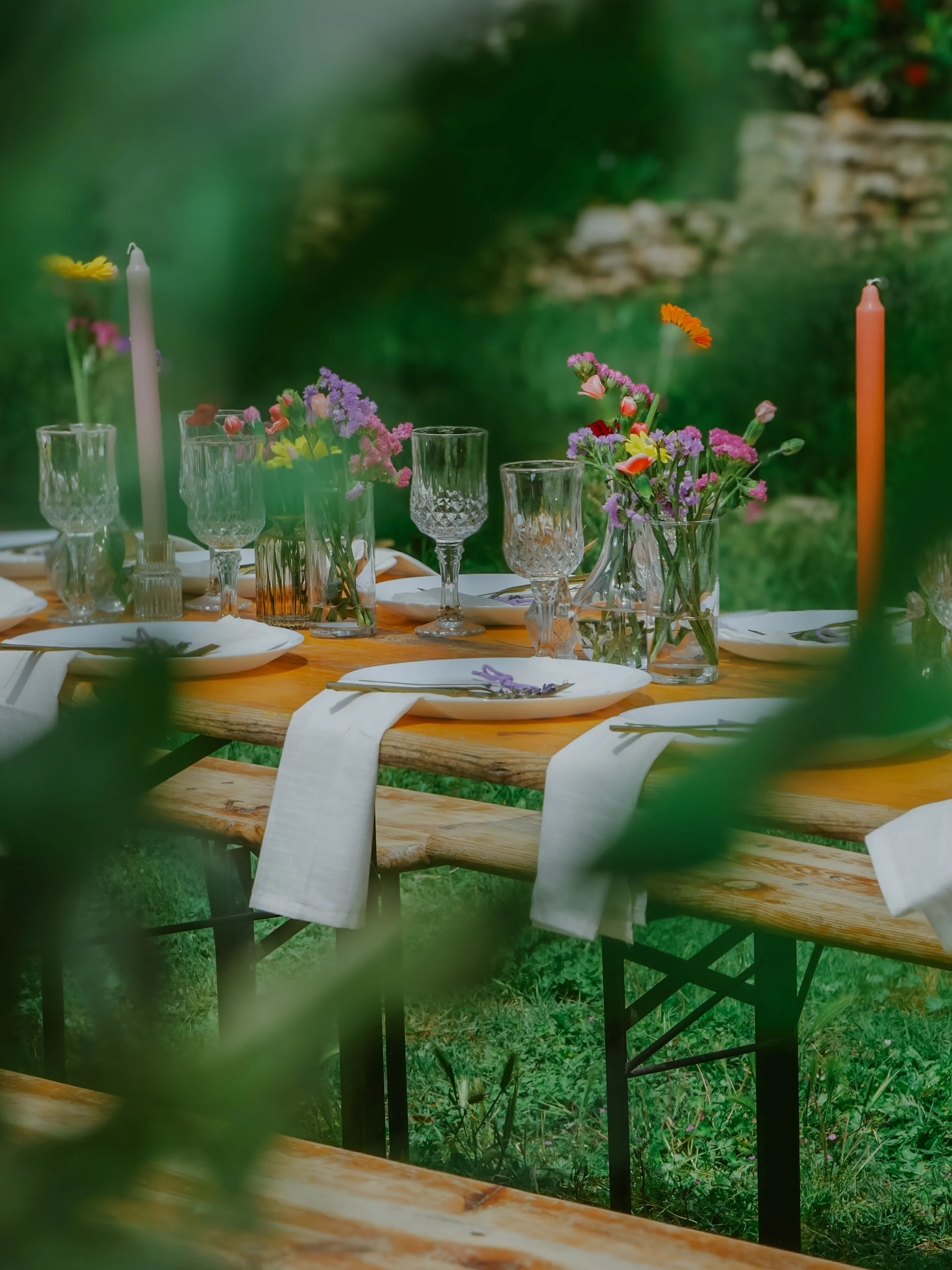 Table en bois rustique décorée avec des vases de fleurs colorées, assiettes blanches, verres en cristal et bougies dans un jardin en pleine nature.