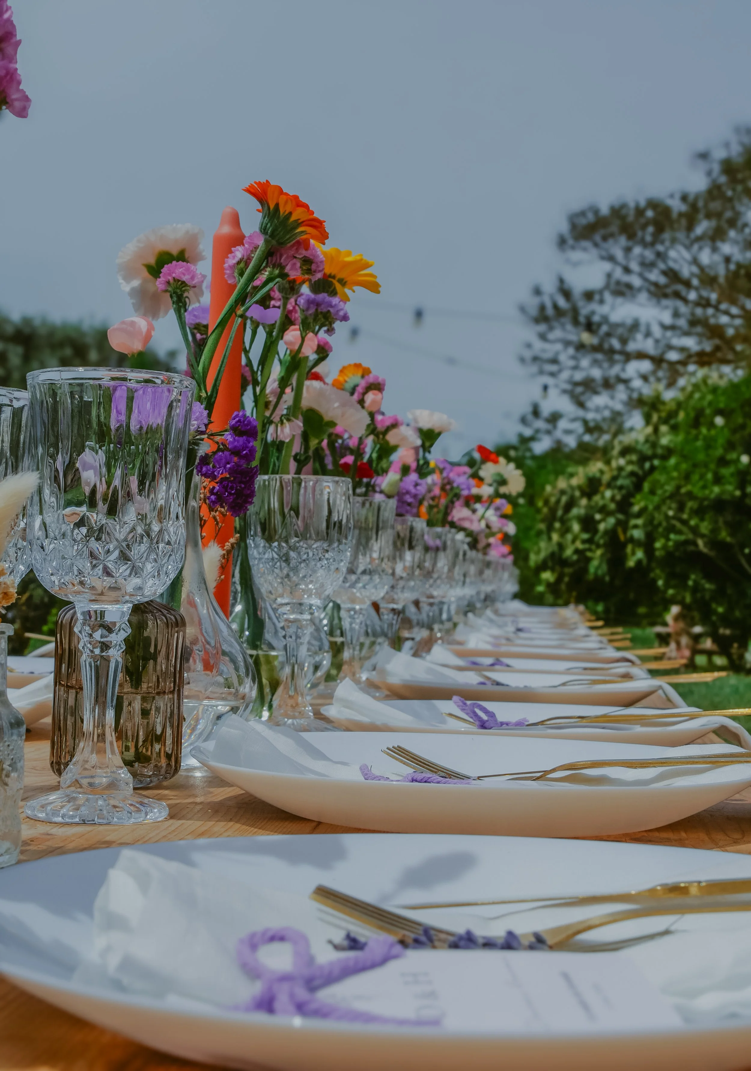 Une table de fête en plein air ornée de fleurs, de verres en cristal et de plateaux avec des nappe blanches, sous un ciel bleu, entourée d'arbres verdoyants.