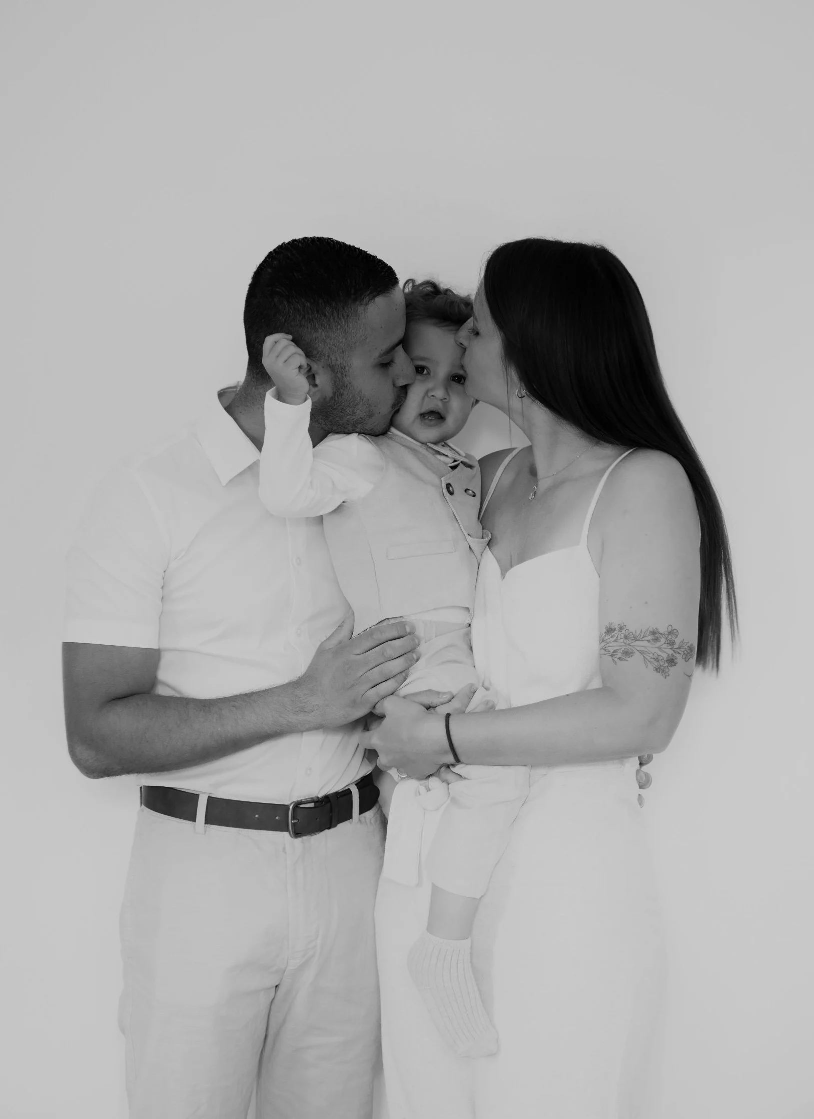 Une famille de trois personnes, un homme, une femme et un enfant, partage un moment affectueux, la femme embrassant l'enfant et l'homme le tenant dans ses bras, tous portant des vêtements clairs.