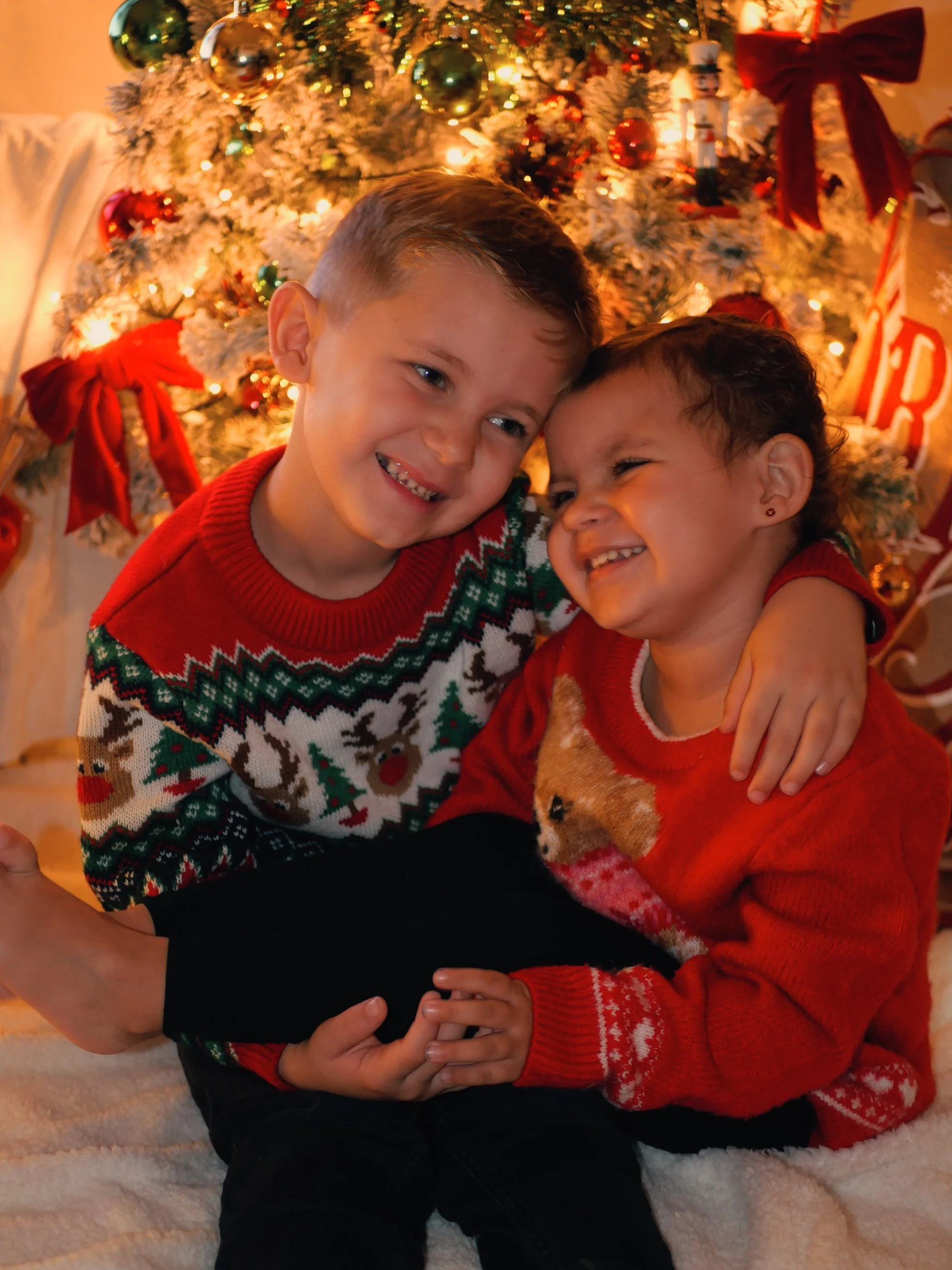 Deux enfants souriants, un garçon avec des cheveux courts et une fille avec des cheveux bouclés, en vêtements de Noël, se font un câlin devant un sapin de Noël décoré de boules et de rubans rouges.