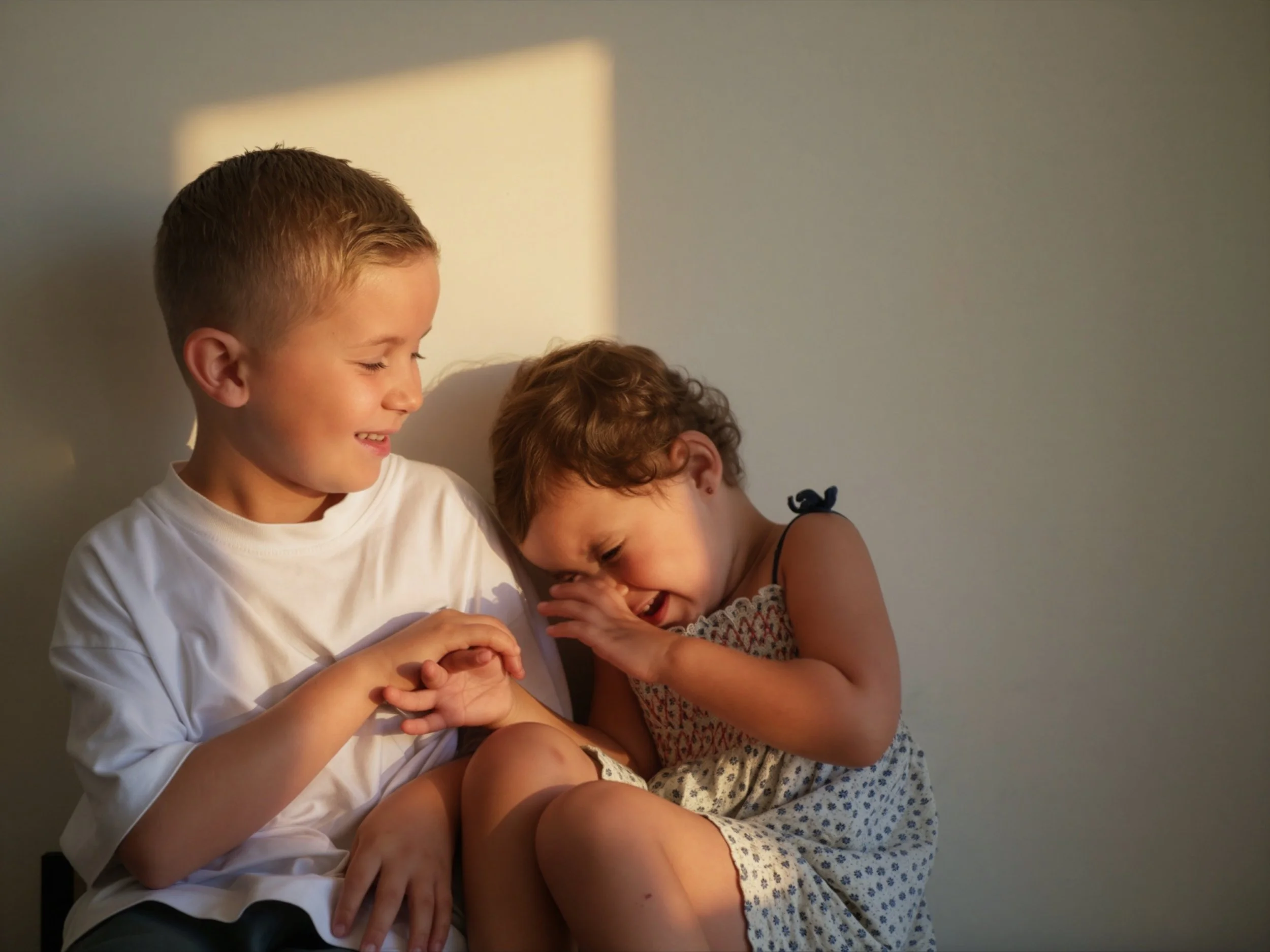 Trois enfants rient et s'amusent ensemble, assis contre un mur beige, une lumière chaleureuse tombe sur eux.