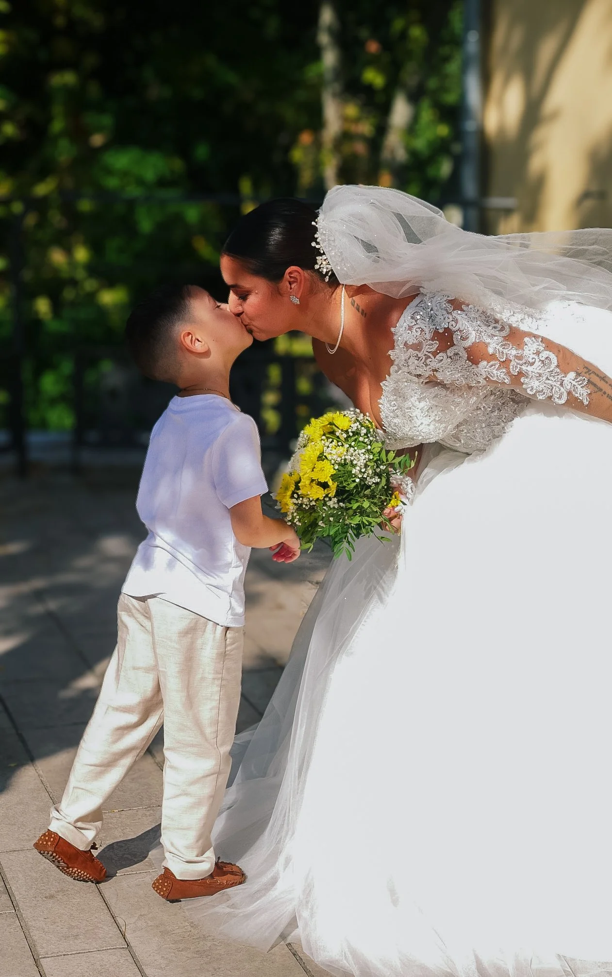 Une femme en robe de mariée embrasse un jeune garçon tenant un bouquet de fleurs, sur une terrasse en plein air.