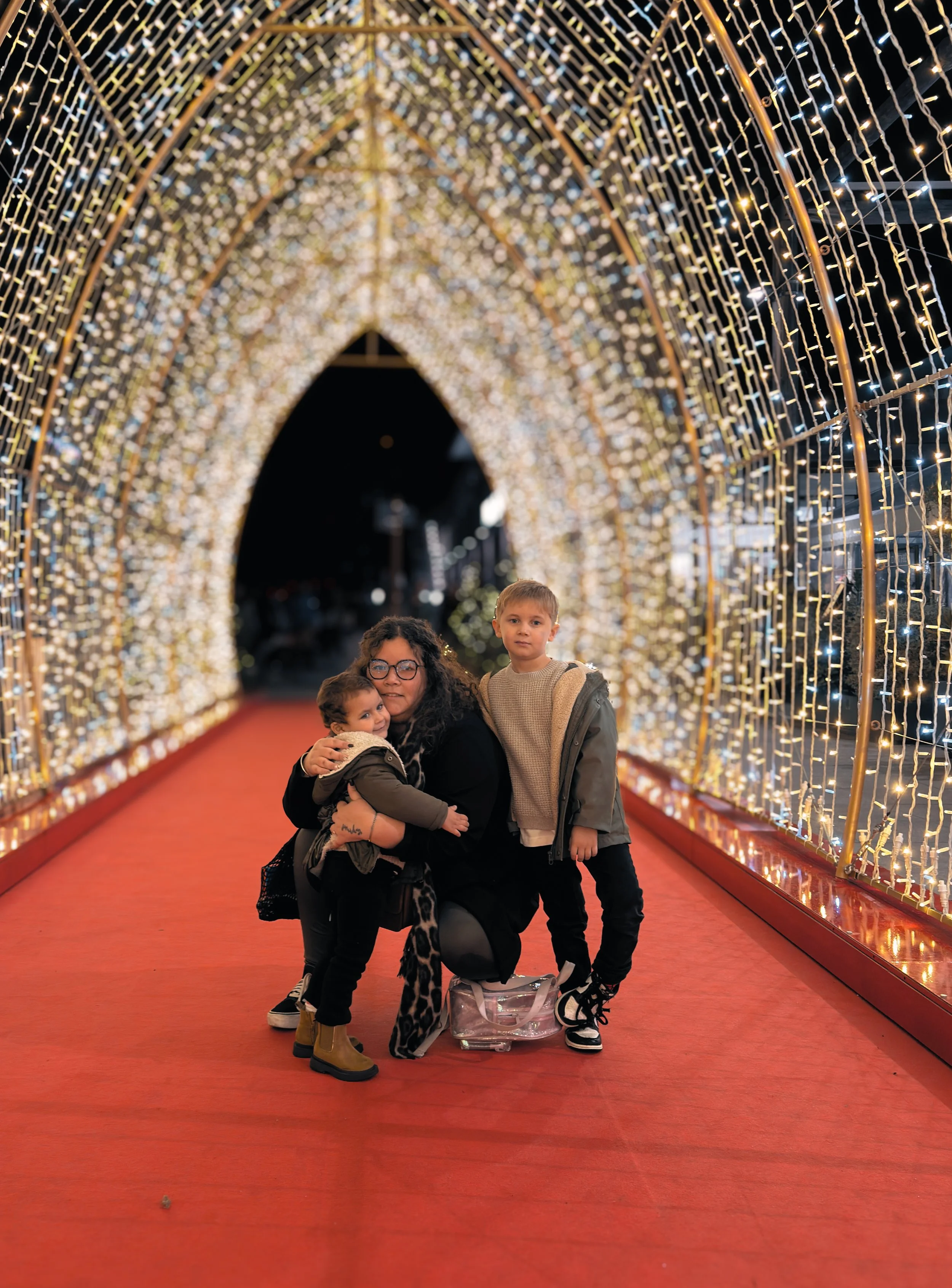 Un groupe de cinq personnes, probablement une famille, posent sous un tunnel décoré de lumières de Noël. La femme au centre tient un bébé, un garçon se tient à côté d'elle, et deux autres enfants sont devant. Le sol est rouge, et l'arche décorative est illuminée.