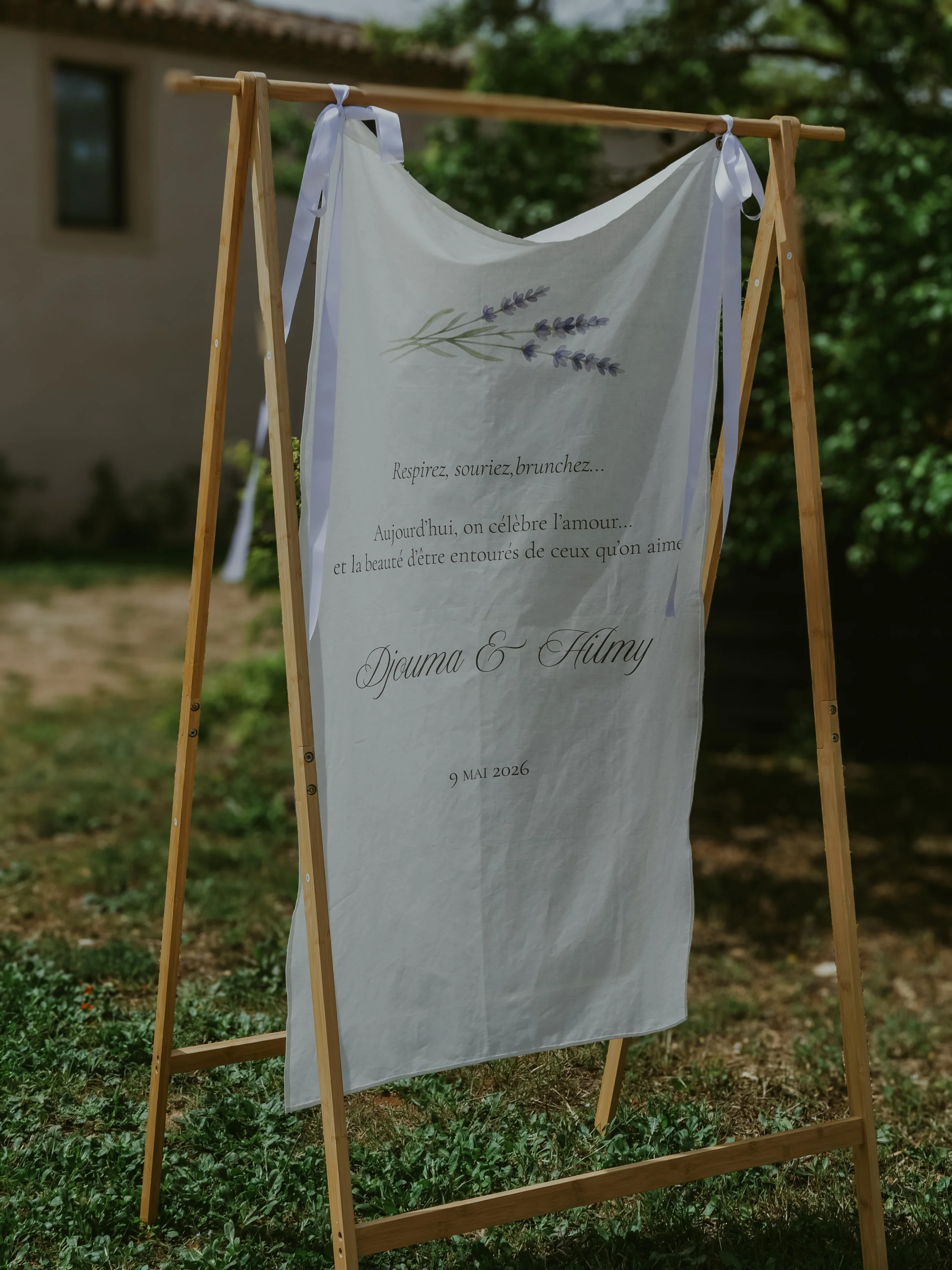 Panneau blanc suspendu sur une scène en bois, avec une illustration de lavande et un message en français. La scène est extérieure, entourée d'arbres verts et d'une maison en arrière-plan.