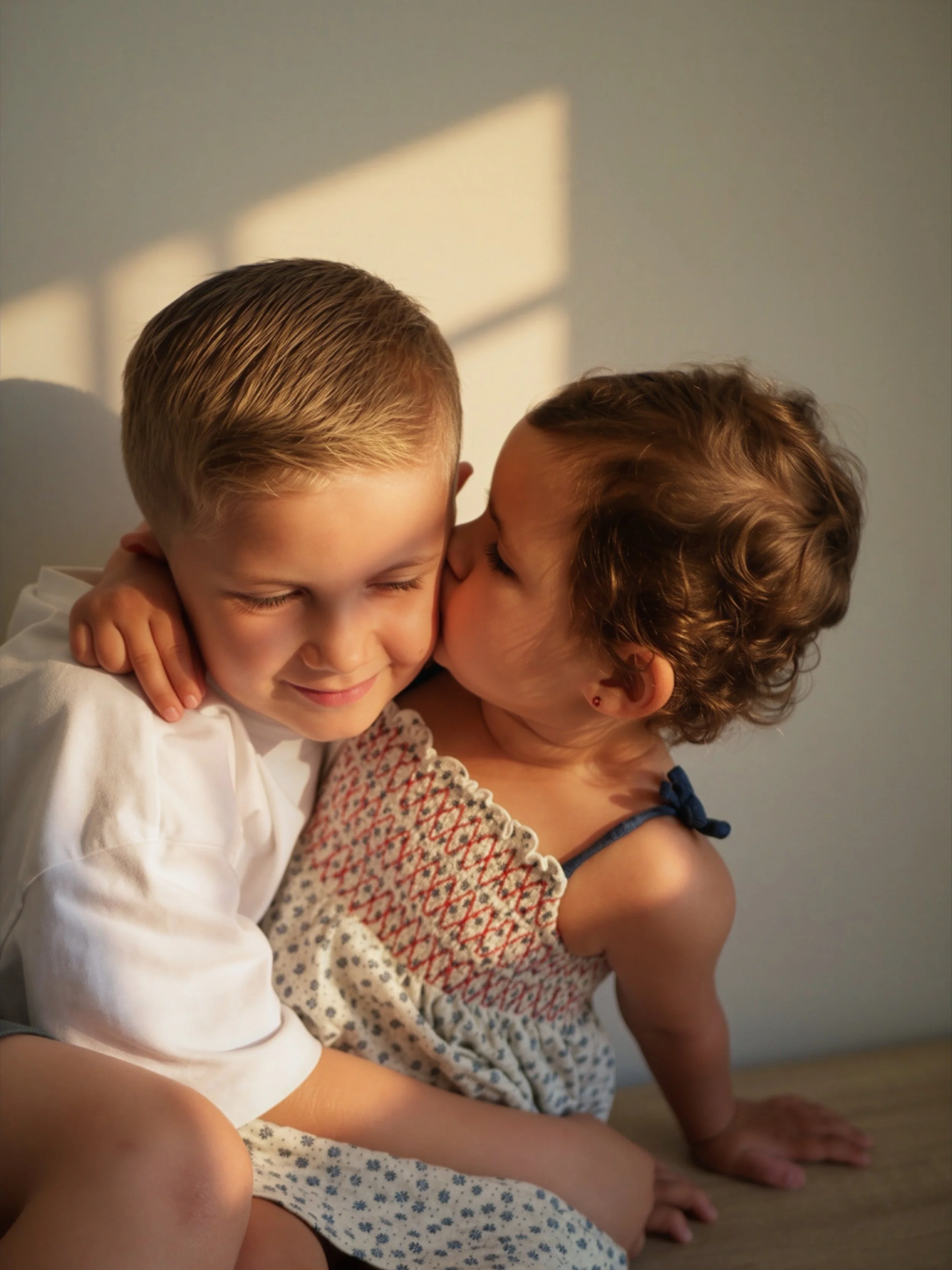 Deux enfants s'embrassent affectueusement, l'un avec les yeux fermés et l'autre qui lui donne un bisou sur la joue, assis par terre à côté d'une mère ou d'une adulte.