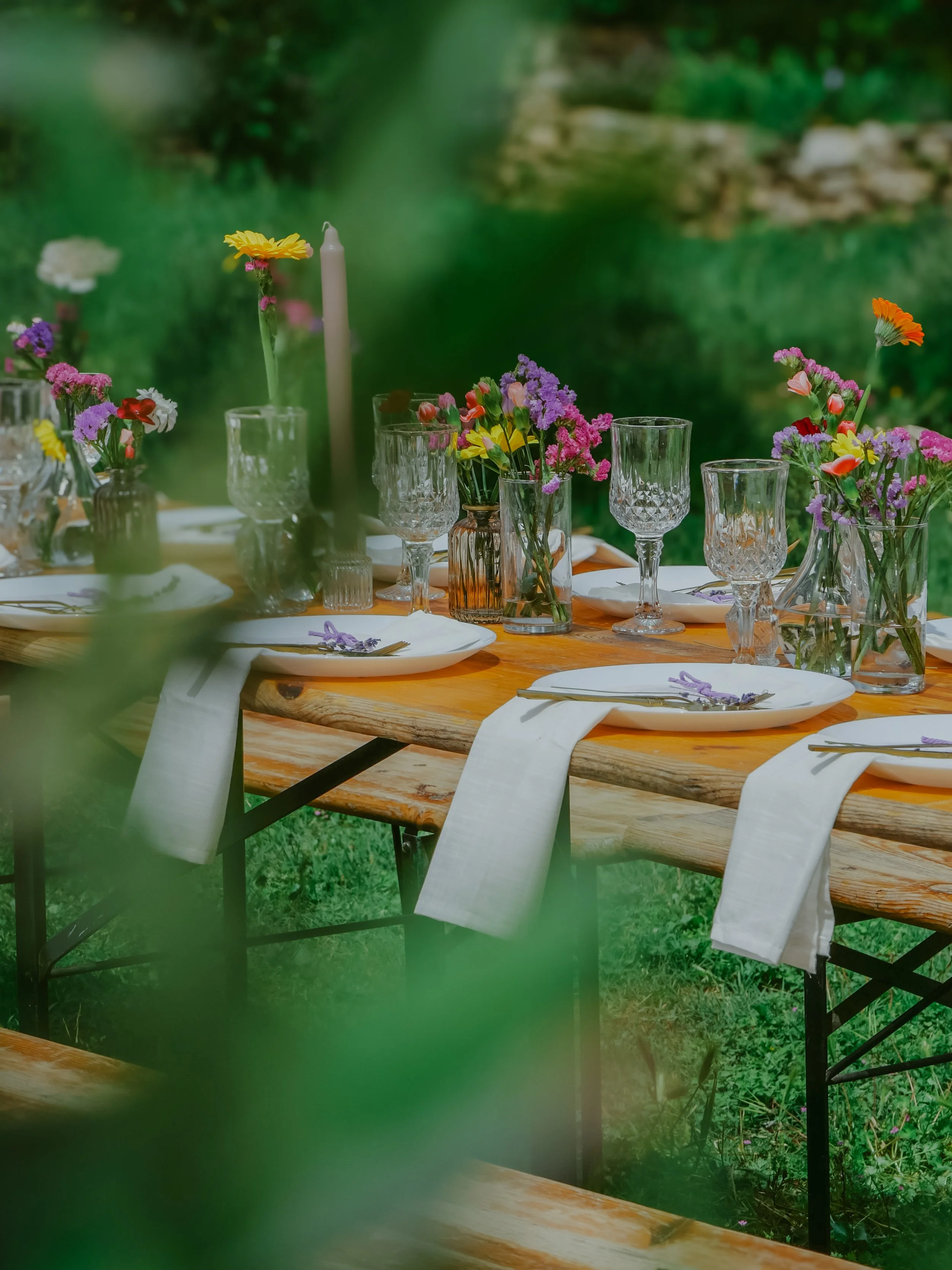 Une table en bois avec des vases remplis de fleurs colorées, des verres à vin, des assiettes blanches et des nappes blanches, située en plein air dans un jardin verdoyant.