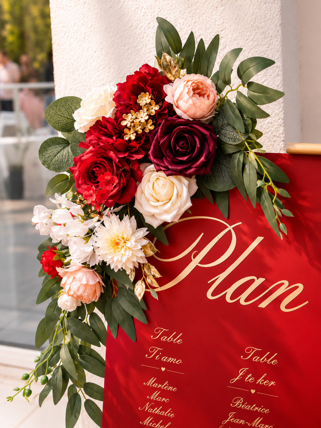 Composition florale avec roses rouges, roses blush, fleurs blanches, feuillage vert foncé et doré, sur un panneau rouge avec écriture en français et italien