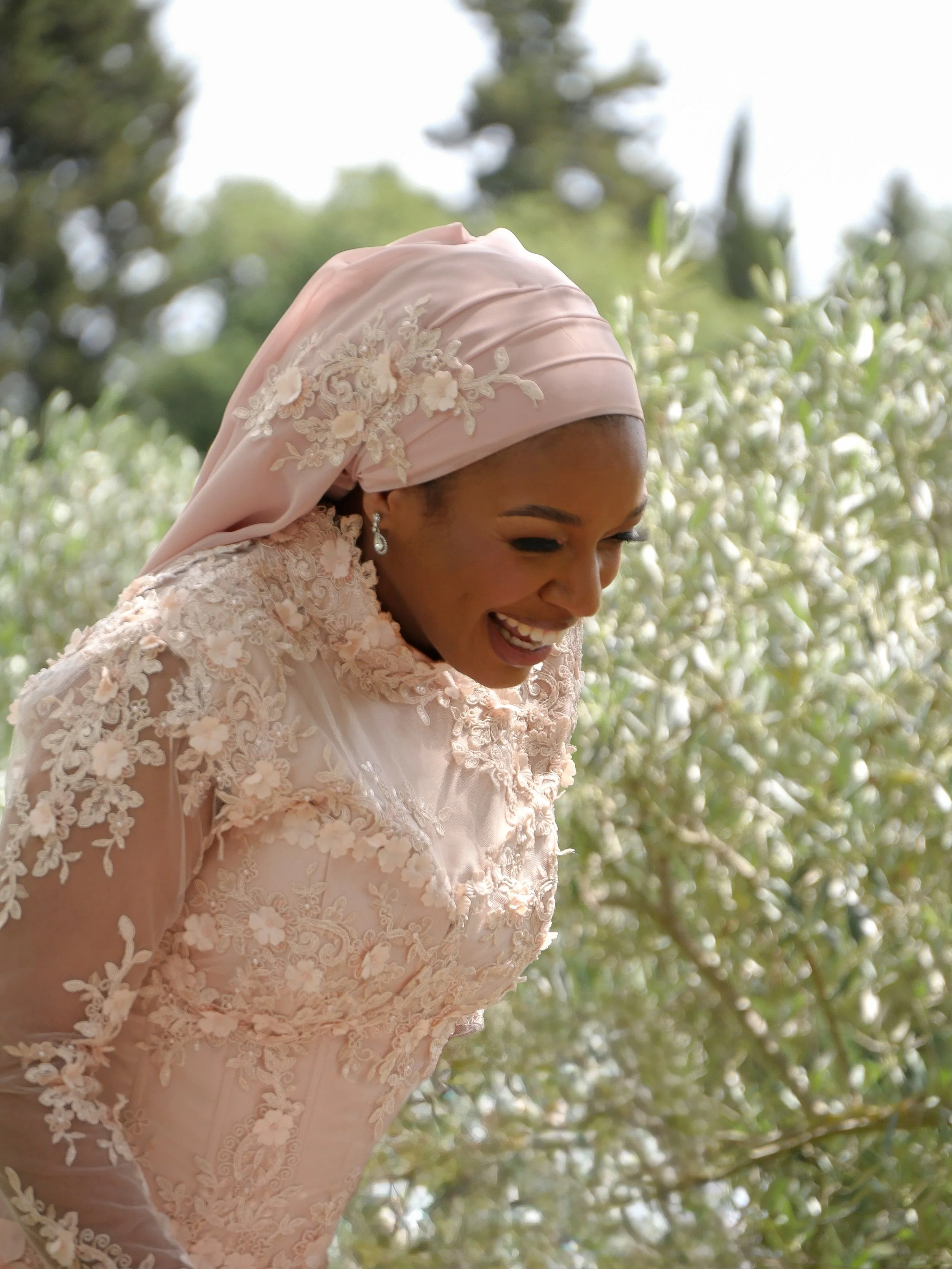 Une femme souriante portant une robe de mariage délicate et un headwrap, entourée de végétation.