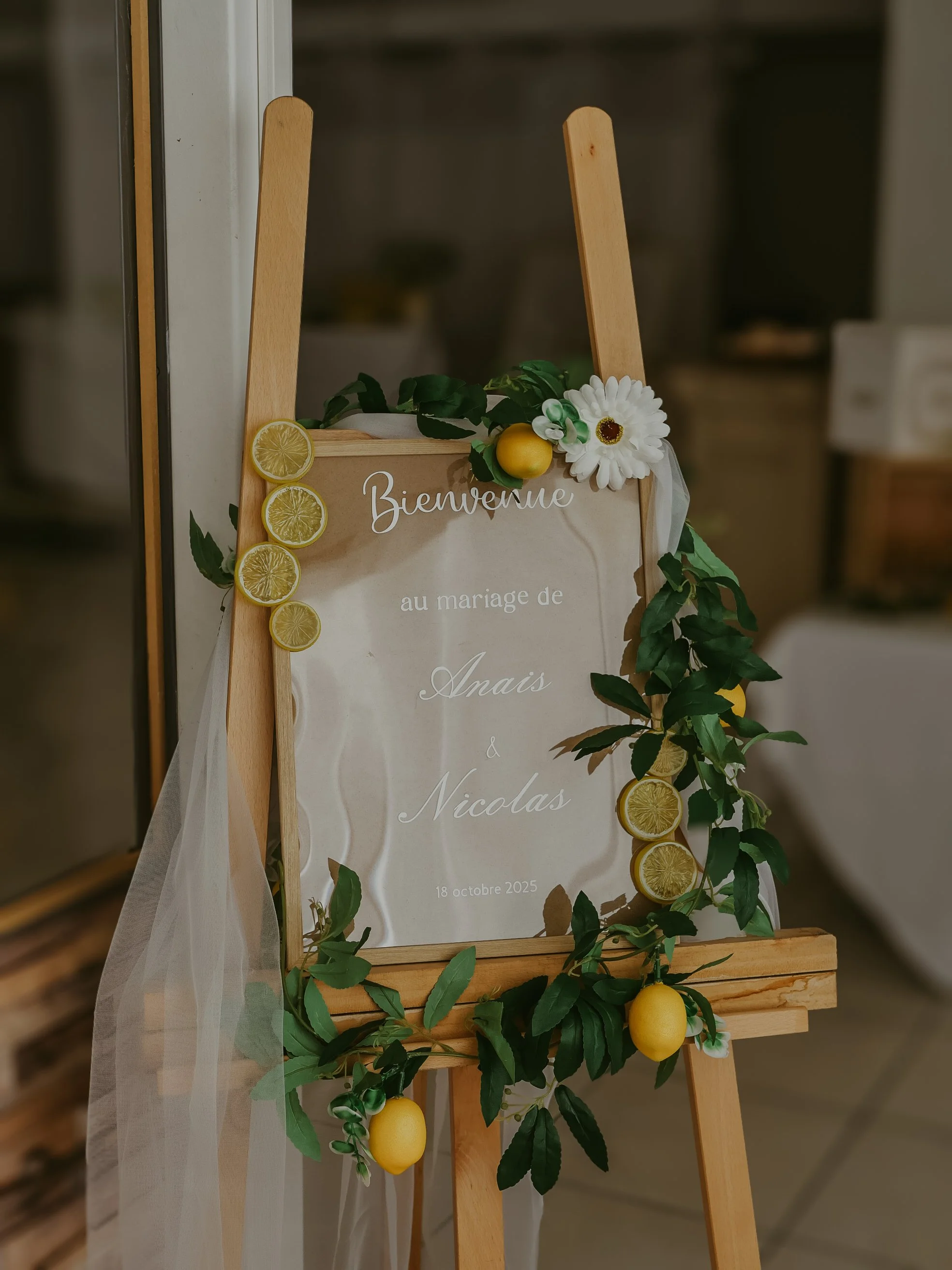 Tableau de mariage décoré avec des citrons, des feuilles vertes, des fleurs blanches, un ruban blanc et un panneau indiquant le mariage de Anaïs et Nicolas, date 18 octobre 2025.