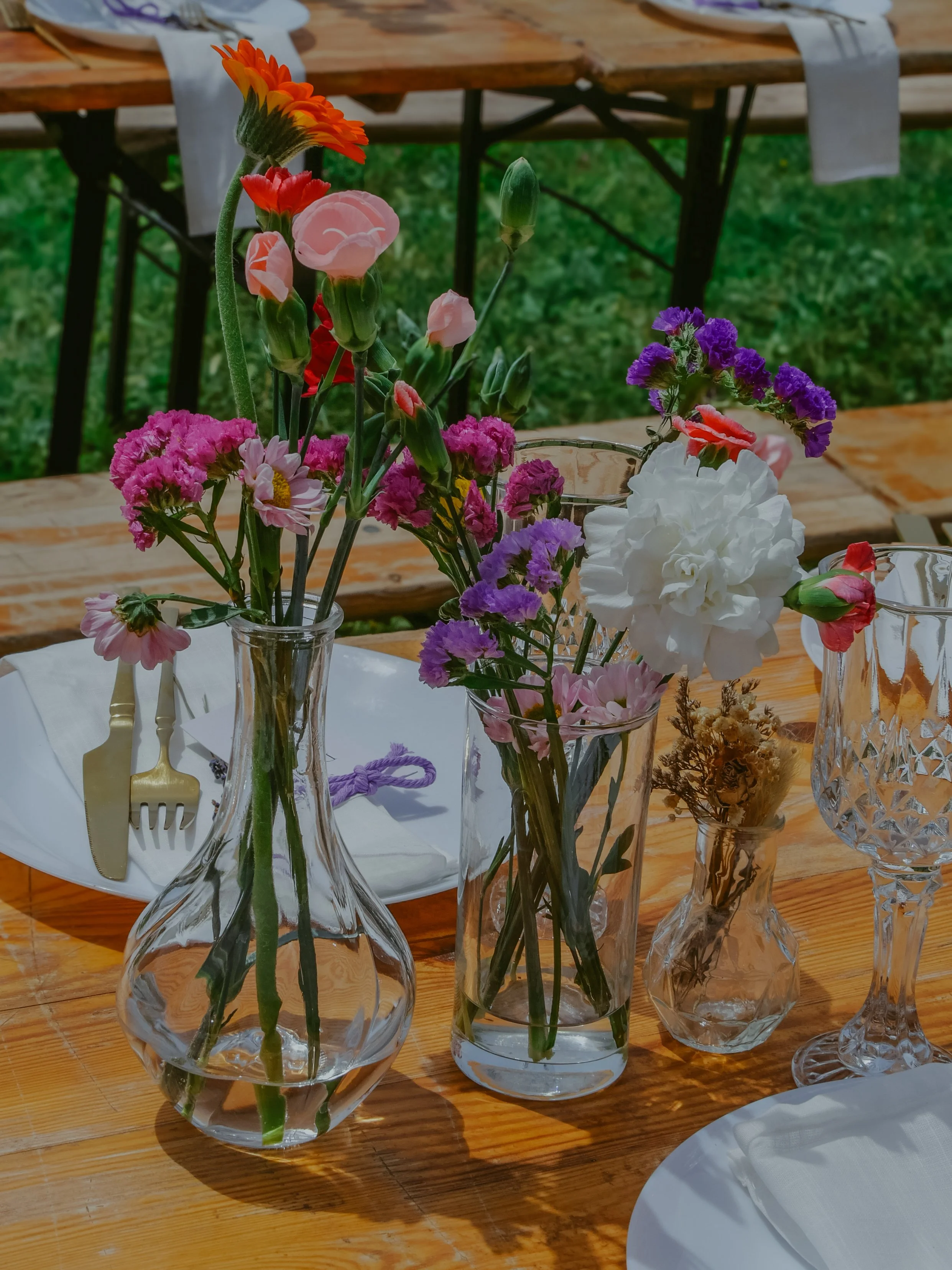 Vases avec des fleurs colorées disposés sur une table en bois lors d'une fête ou d'un repas en plein air.