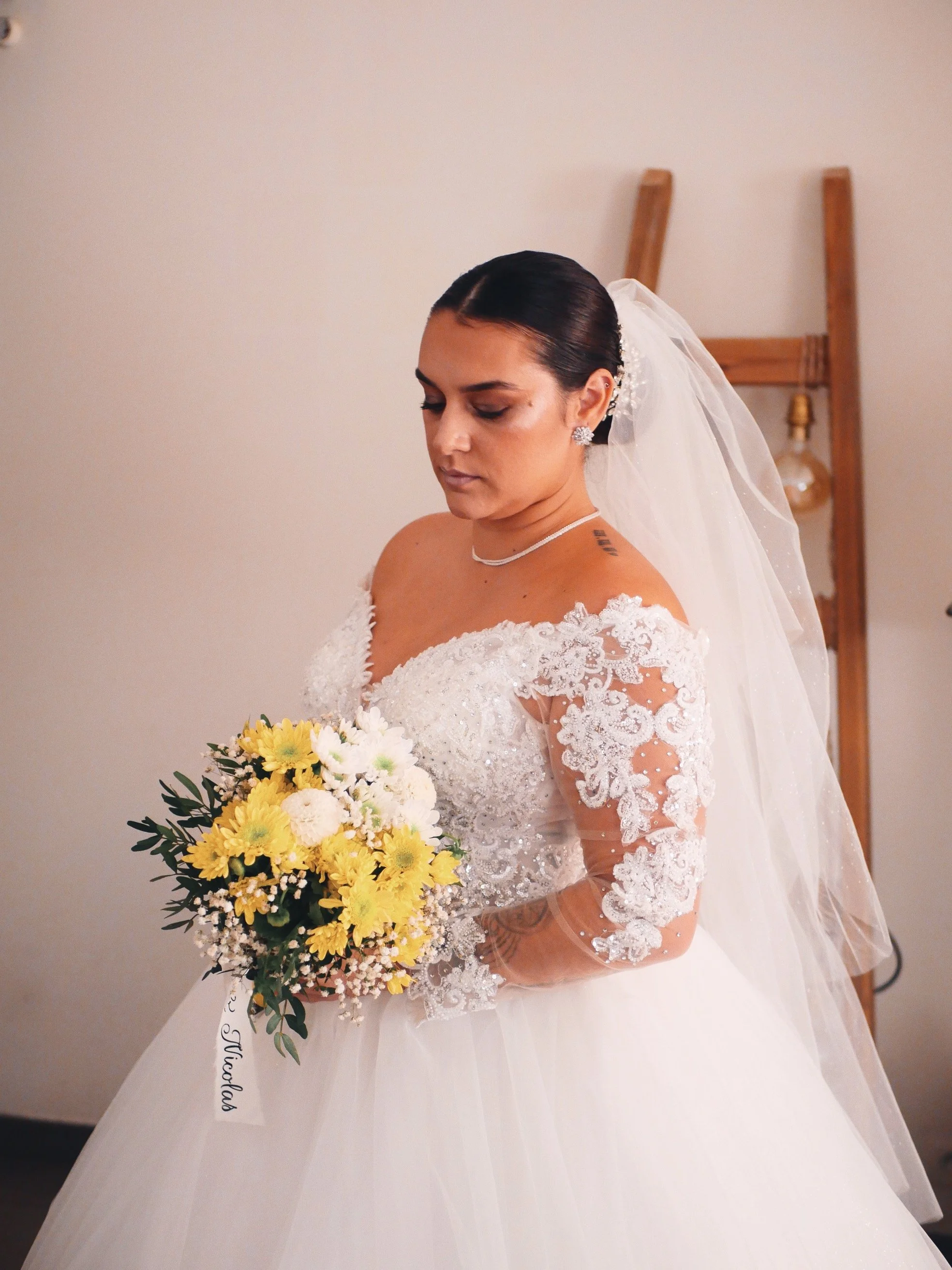 Une femme en robe de mariage blanche avec un voile, tenant un bouquet de fleurs jaunes et blanches, regardant vers le bas, devant un mur blanc, avec des chaises en bois derrière elle.