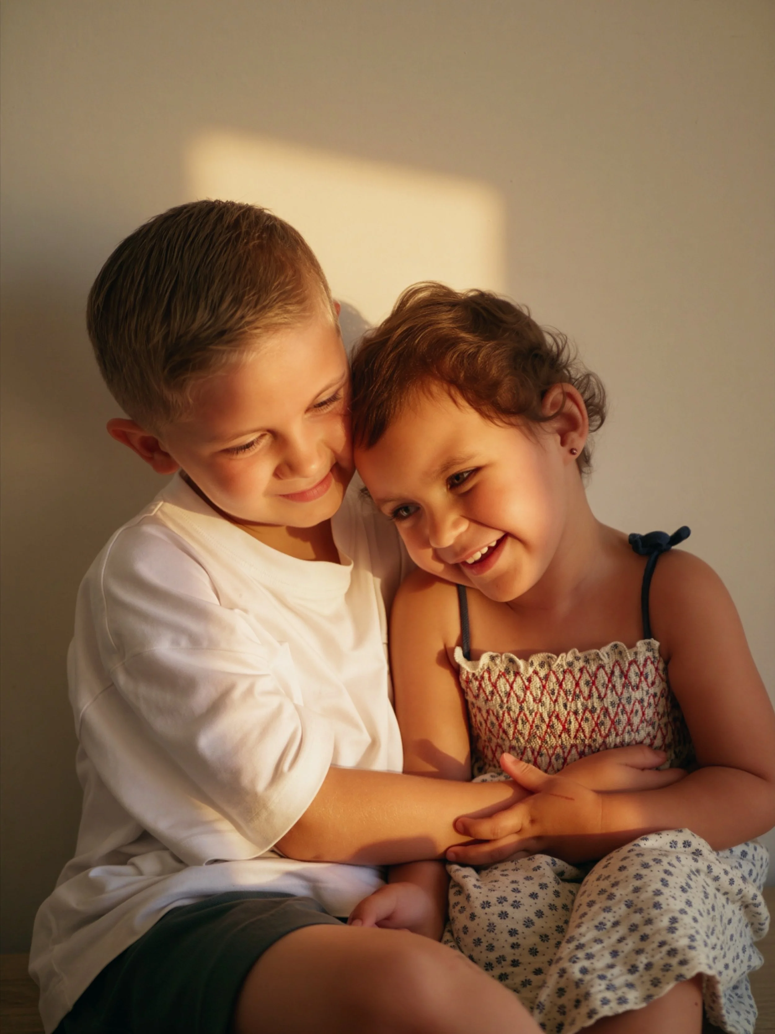 Deux enfants souriants et proches, partageant un moment heureux, assis ensemble contre un mur beige.