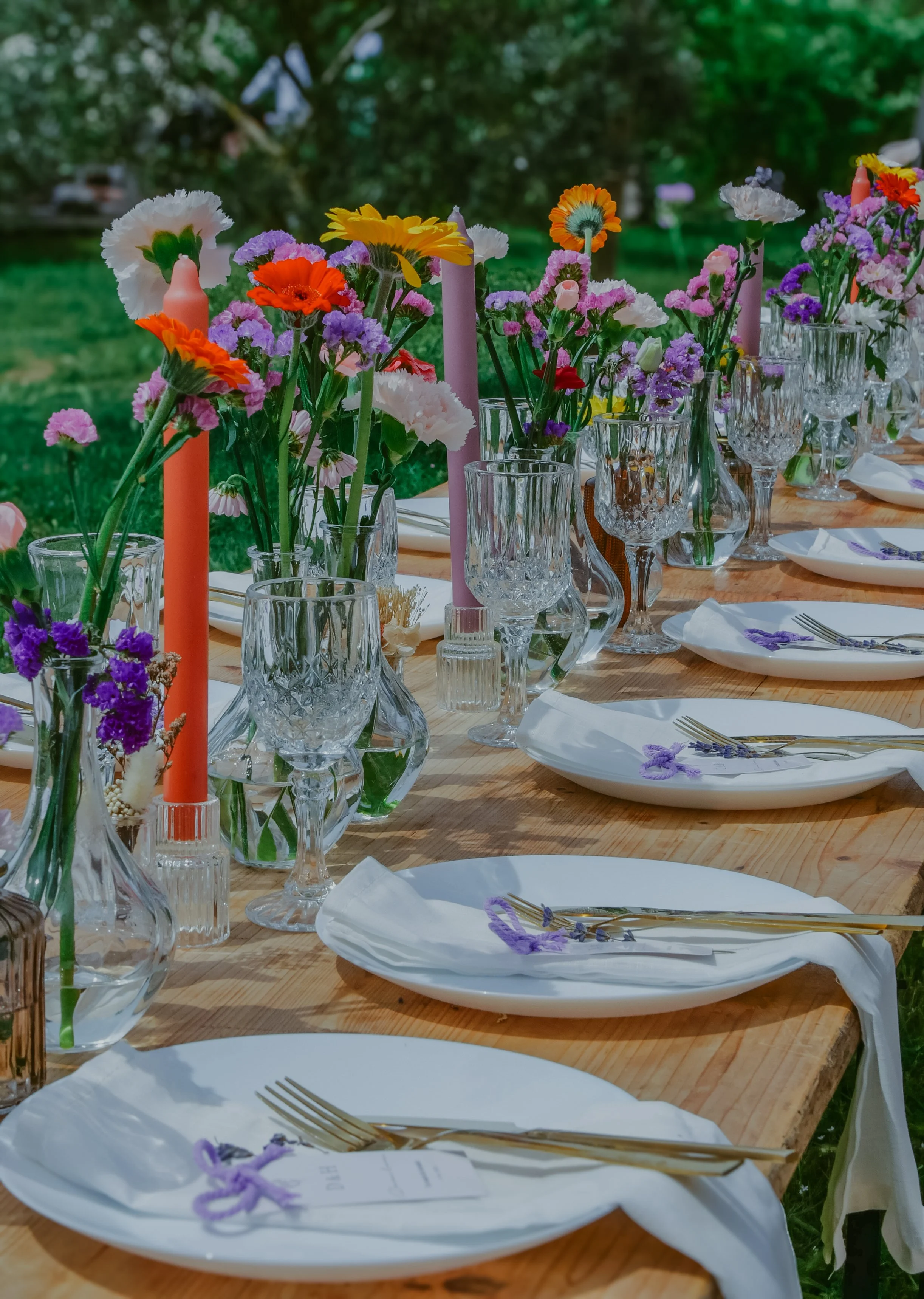 Une table dressée avec des fleurs colorées en vases en verre, des bougies, des assiettes blanches avec des fourchettes, et des nappes blanches, en extérieur dans un jardin verdoyant.
