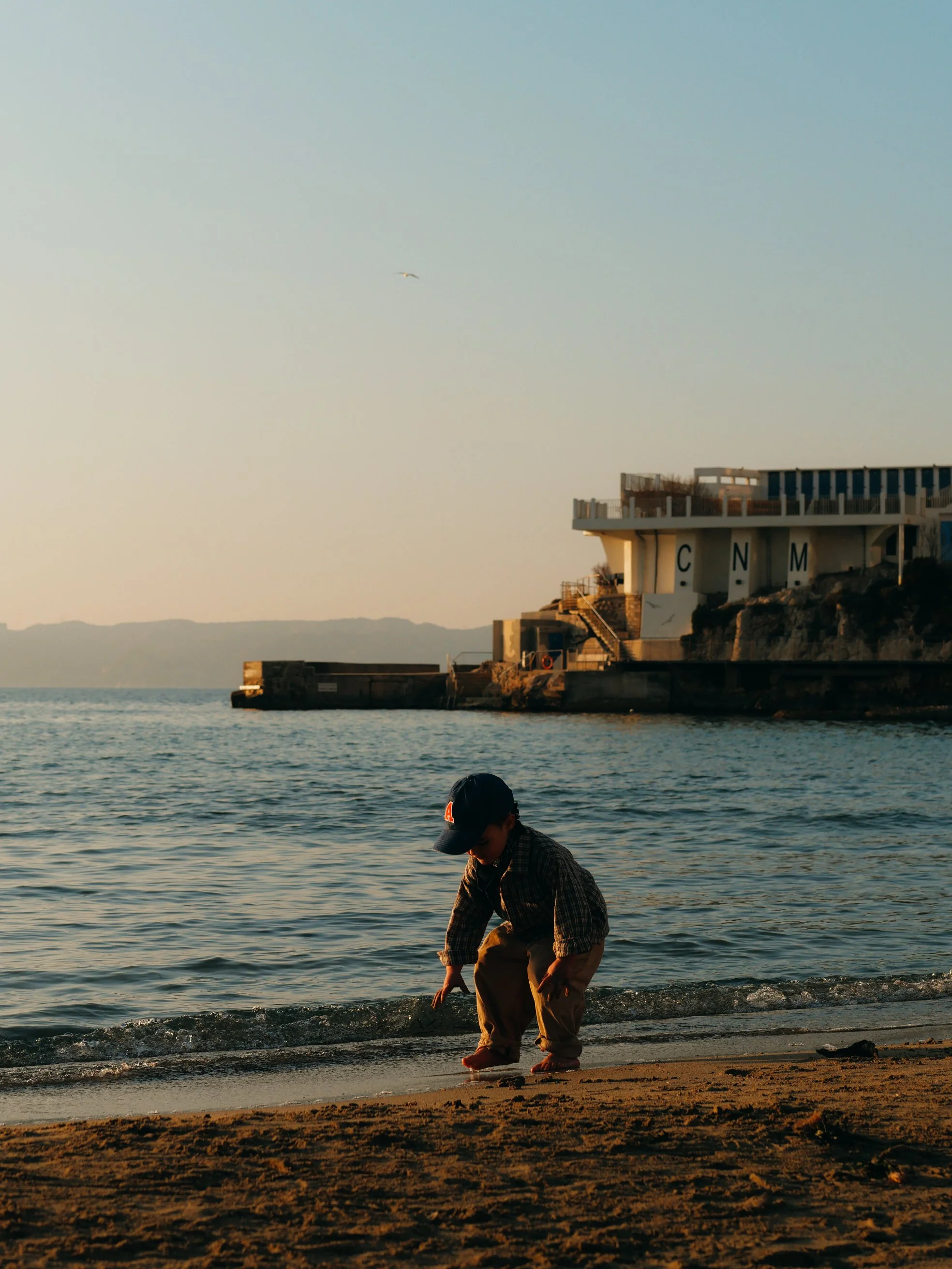 Un jeune garçon en vêtements décontractés joue au bord de la mer, avec un bâtiment en arrière-plan et un oiseau dans le ciel au coucher du soleil.