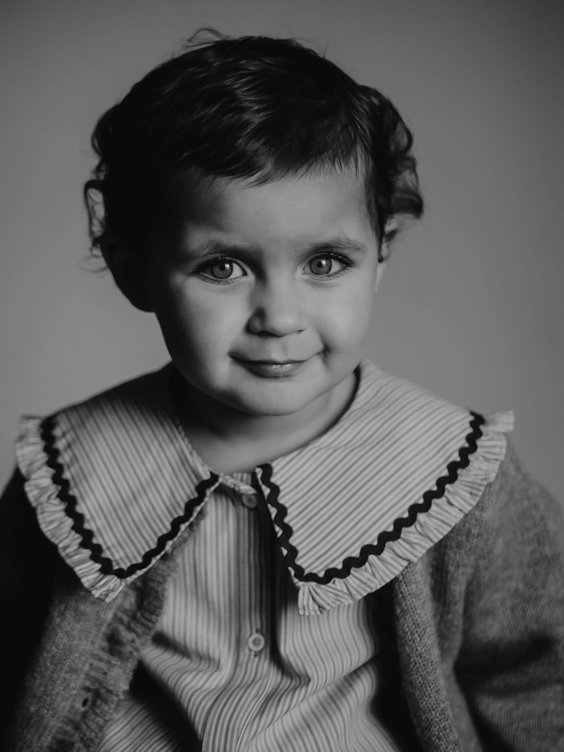 Portrait en noir et blanc d'une jeune enfant avec des cheveux courts et bouclés, portant un col à rayures avec des détails zigzag, souriant légèrement.