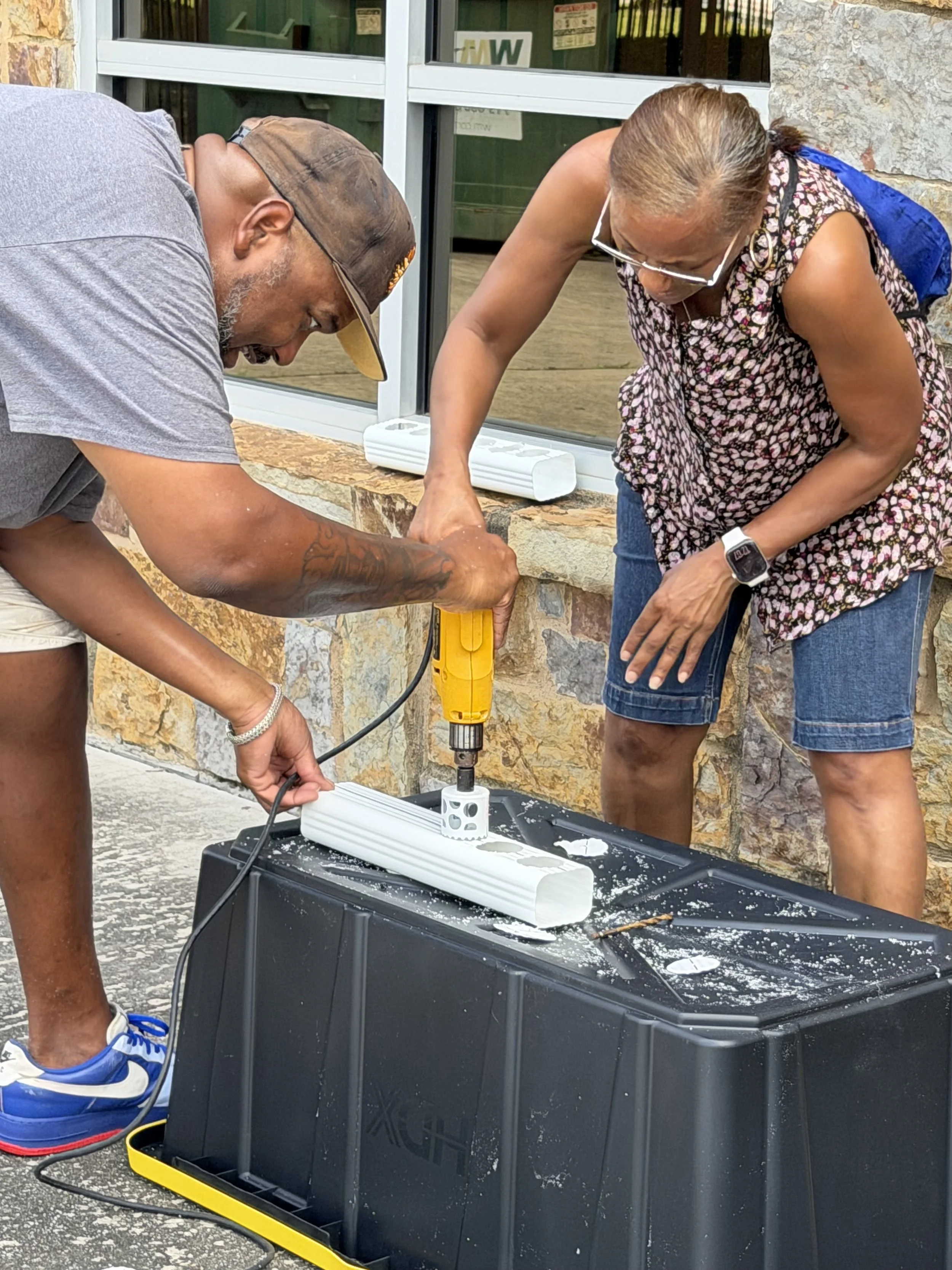 Two people working together outside on a plumbing or electrical project, using a power tool on a white conduit or pipe, with a black plastic container and a stone wall in the background.