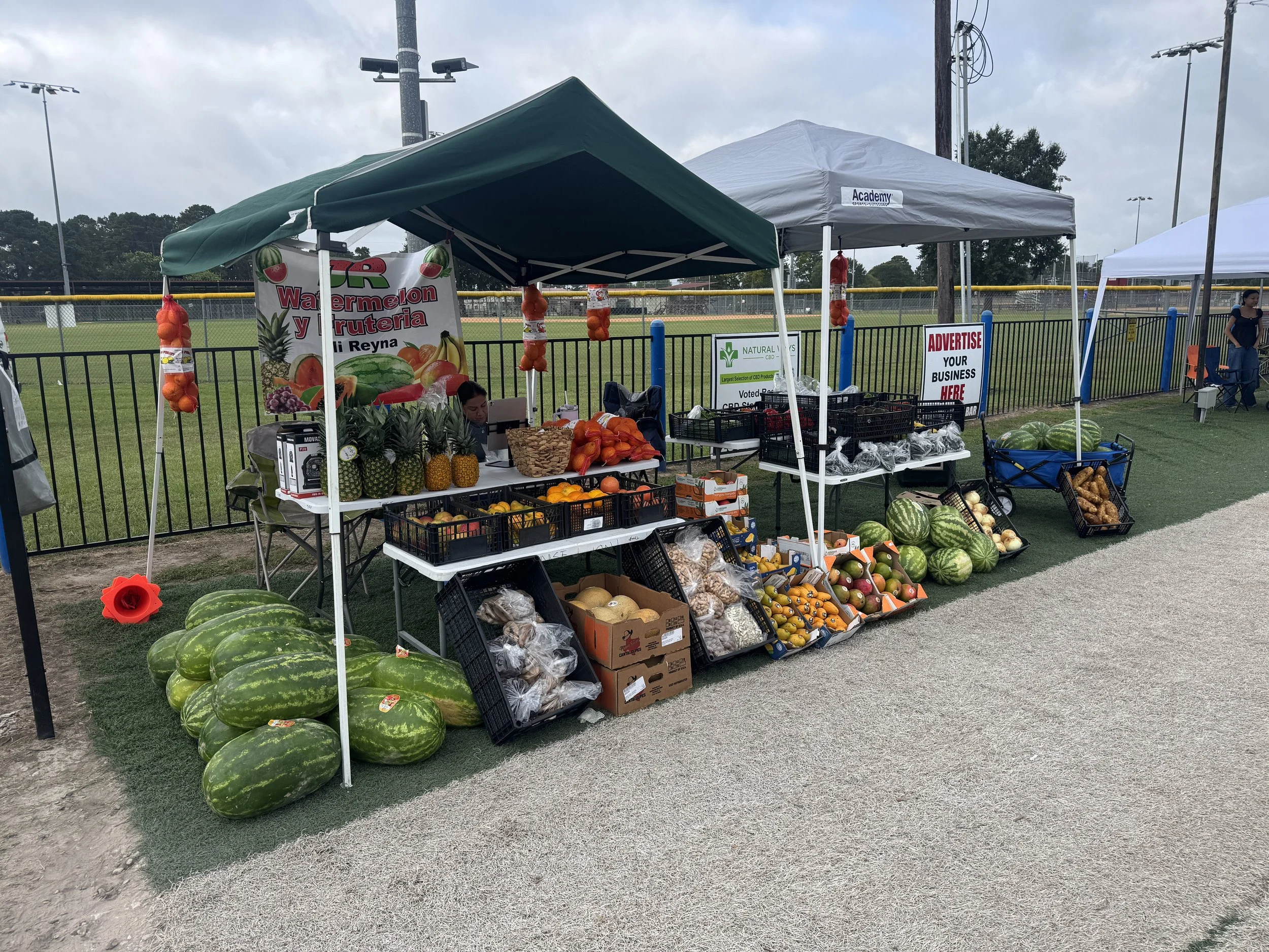 Outdoor farmer's market stand selling watermelons, pineapples, and other fruits, with tents and a baseball field in the background.