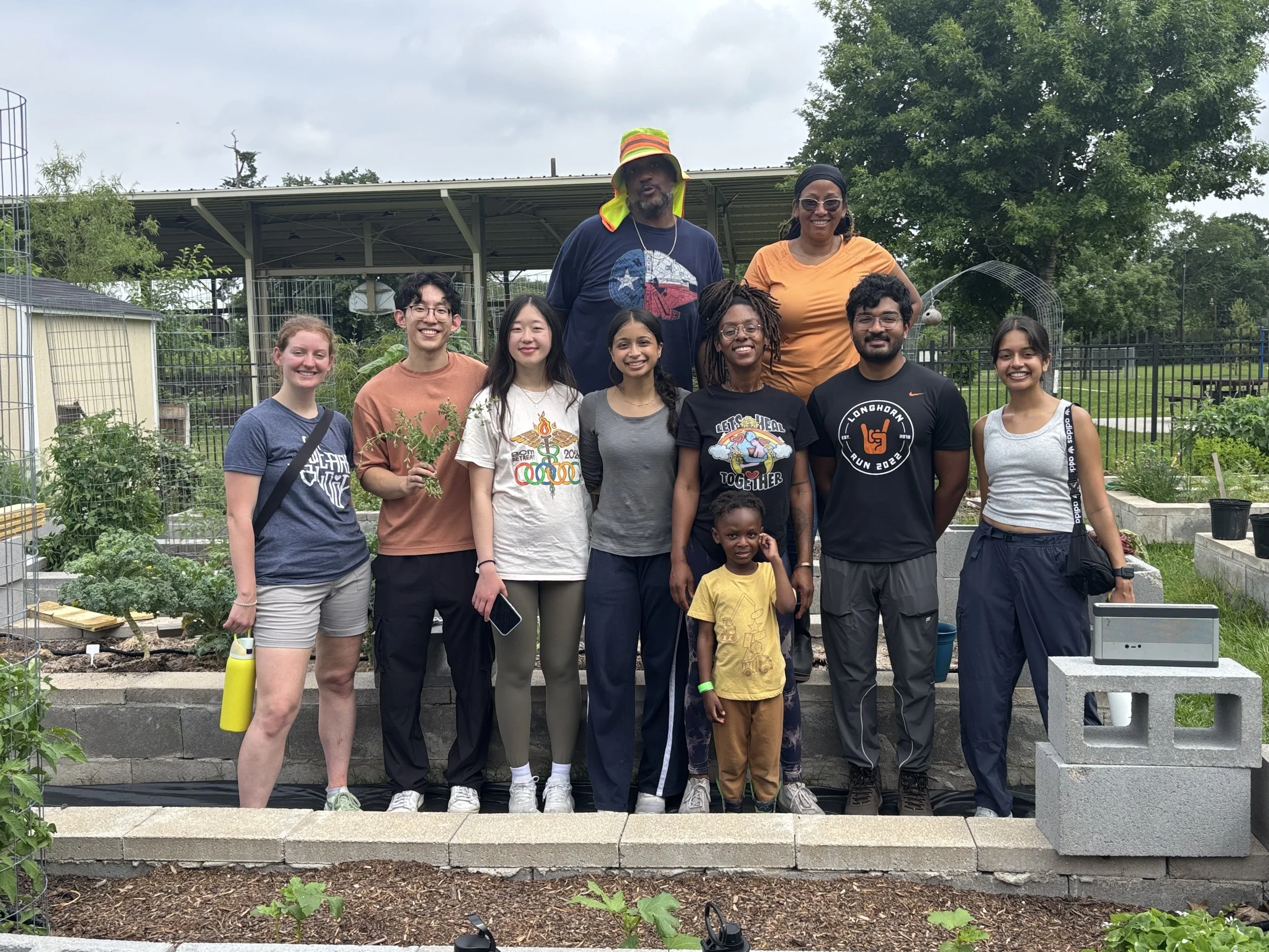 Group of nine diverse people, including children, posing in a community garden with raised beds and trees in the background.