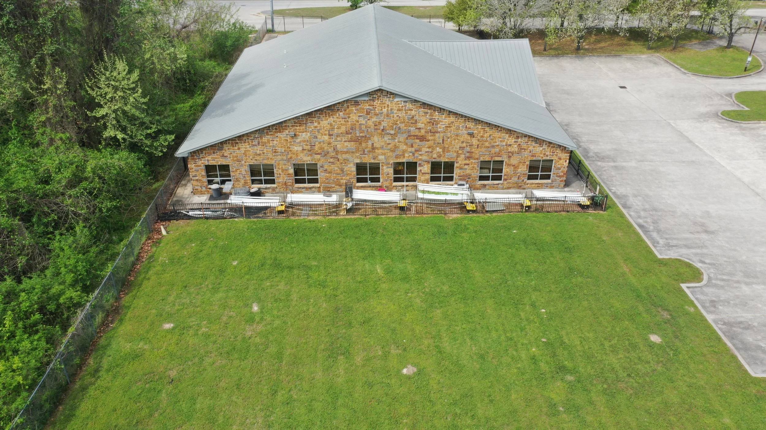 An aerial view of a brick building with a metal roof, surrounded by green grass and trees, with a parking lot nearby.