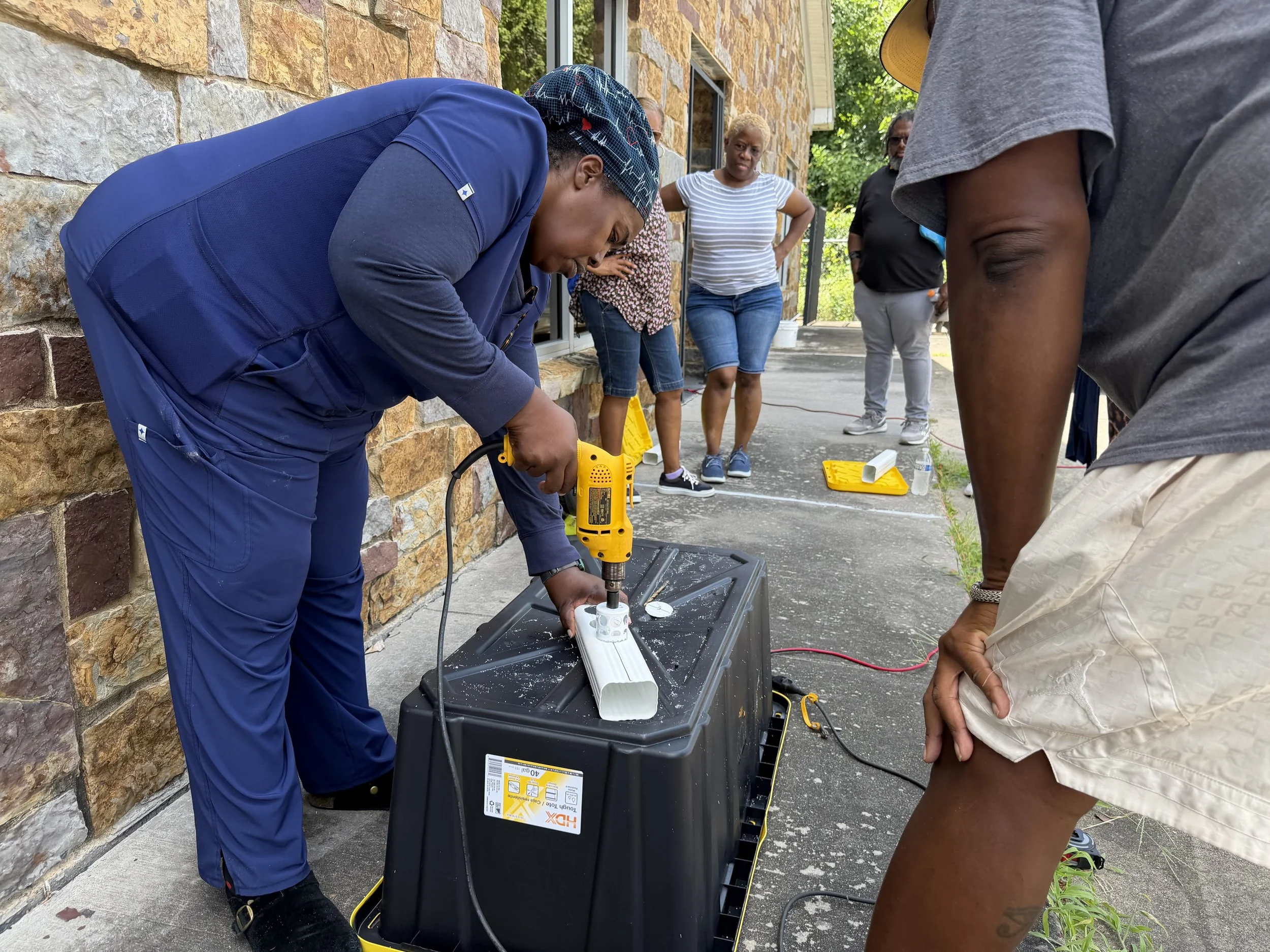 Group of people gathered outside around a person using a drill on a white pipe.