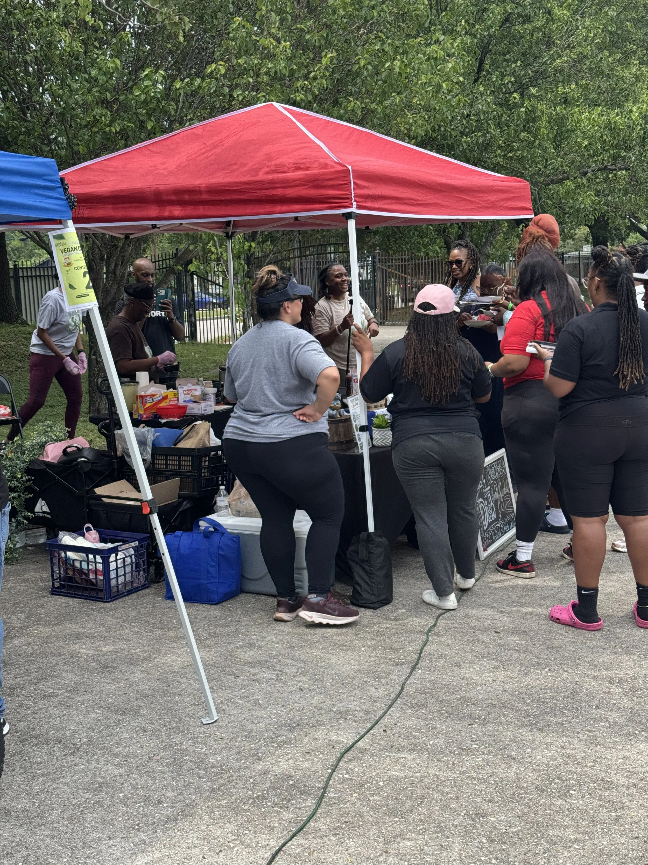 A group of people gathered under a red canopy tent outdoors, engaging in conversation and activities. There are tables with various items, and some people are taking photos or using their phones. The scene is set near a green, leafy area with a fence in the background.