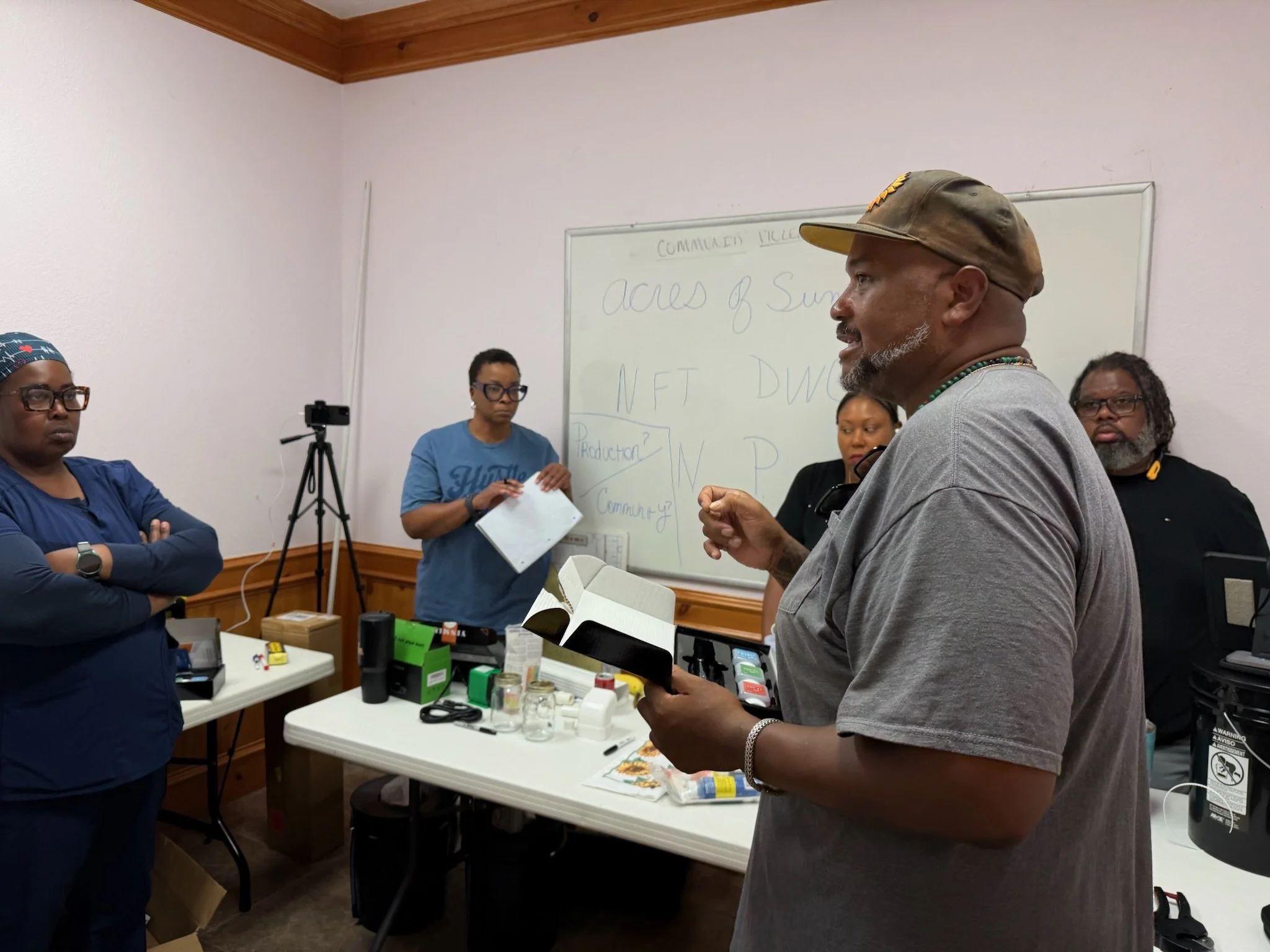 A group of people in a room with one person speaking and others listening, with a whiteboard in the background that has writing related to community and NFTs, and various items like paper, tape, and small containers on a table.