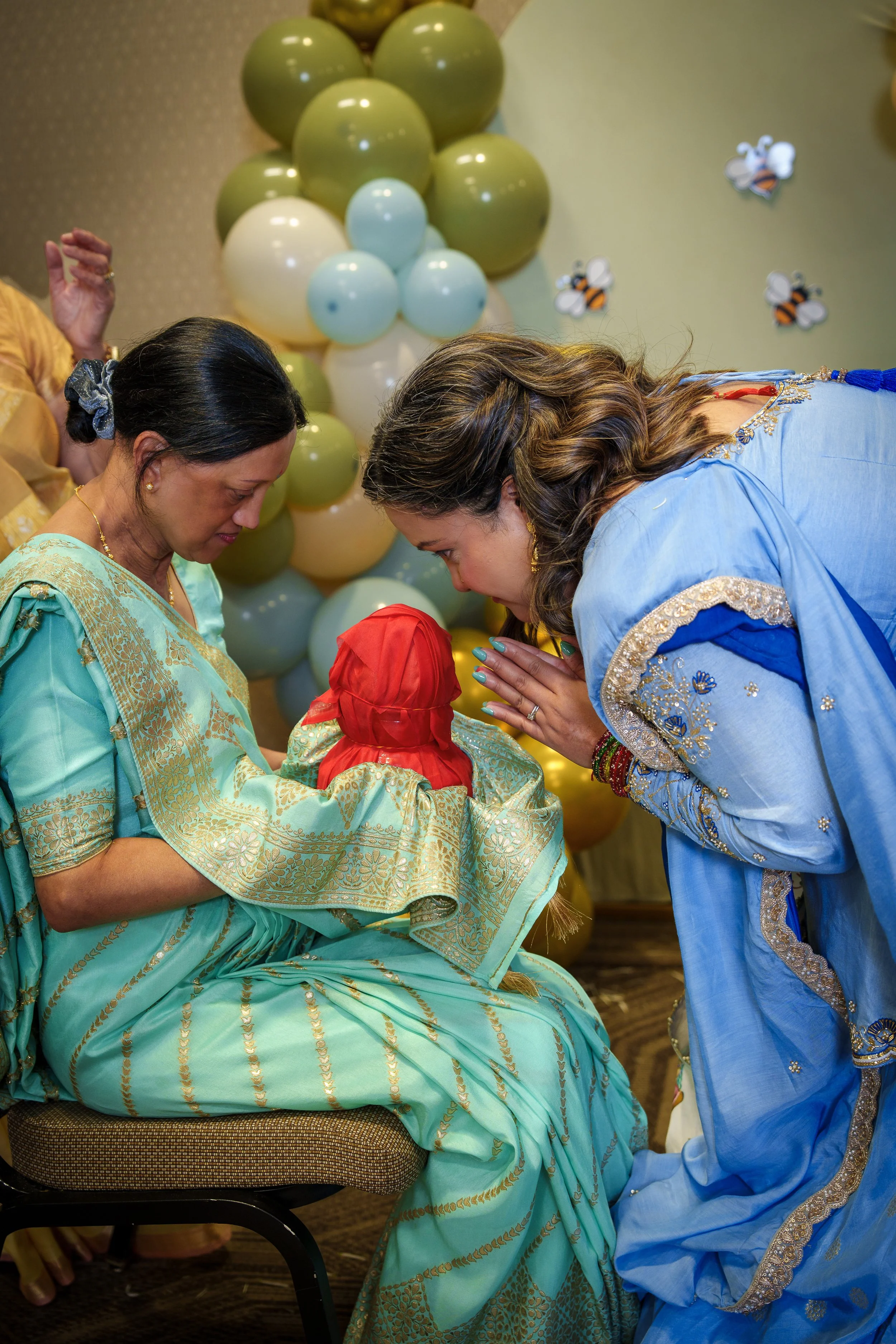 An older woman in a green and gold sari holds a red-wrapped bundle in her lap while a younger woman in blue bows toward her, hands pressed together, foreheads nearly touching.