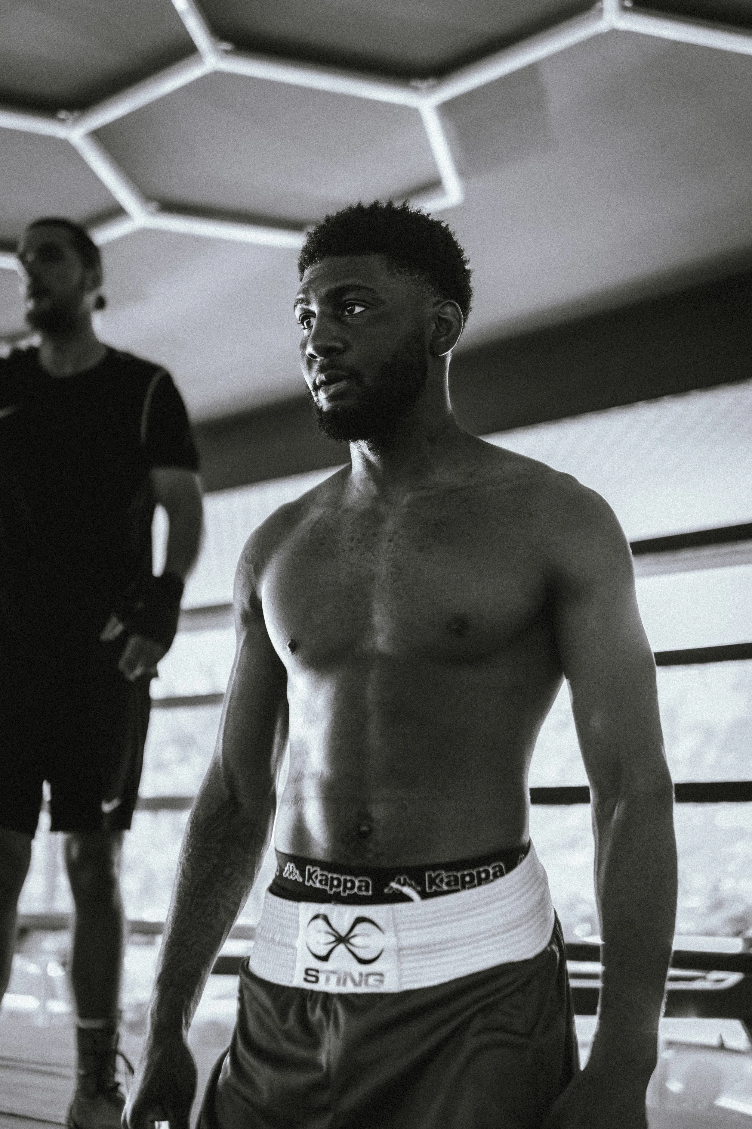 Schwarz-Weiß-Foto eines jungen Mannes ohne Oberbekleidung, der in einem Ring steht, mit einem anderen Mann im Hintergrund, in einem Motiv des Box-Trainings.