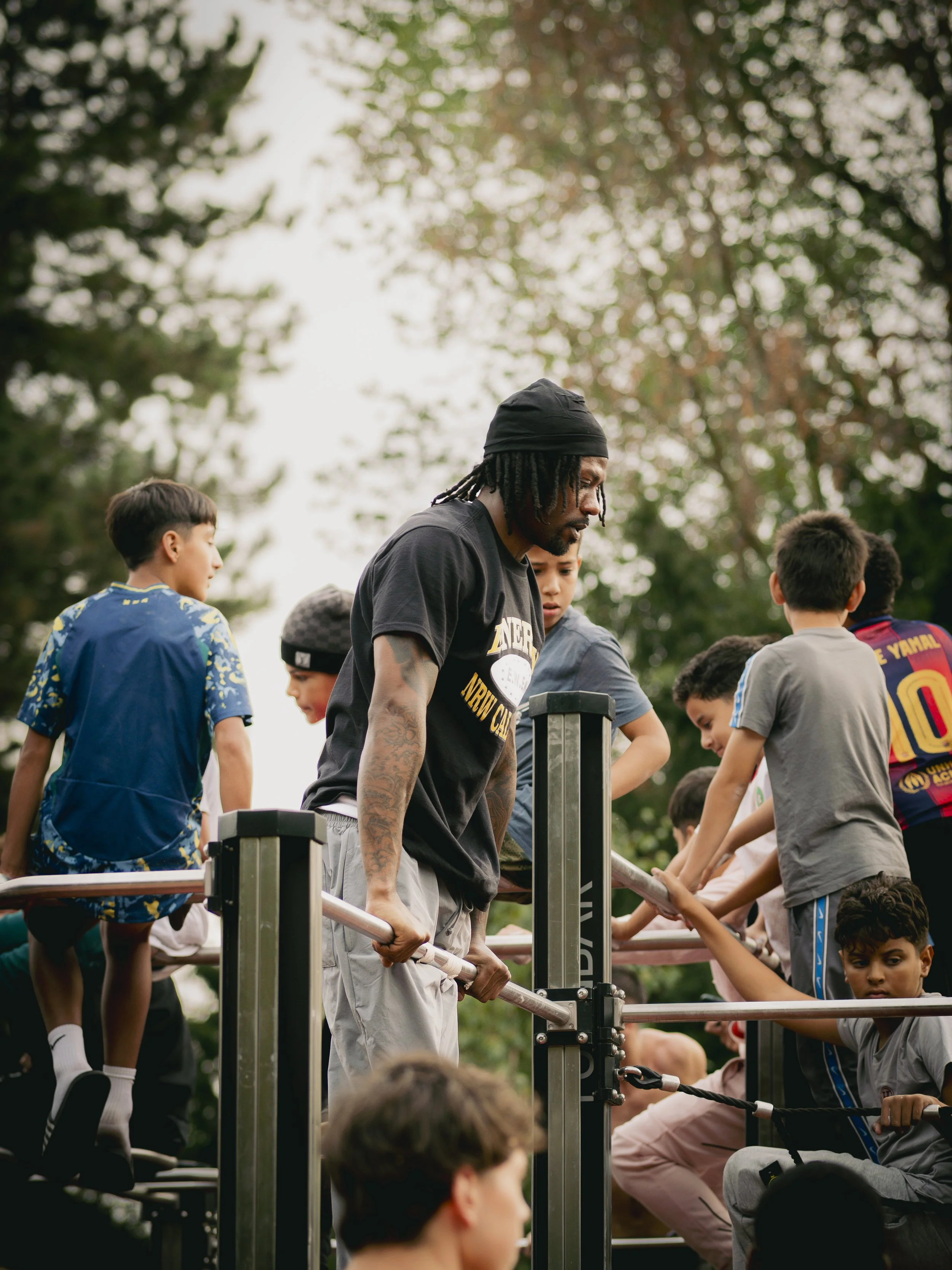 Ein Erwachsener mit dunkler Hautfarbe und Dreadlocks, der eine Gruppe junger Kinder beim Calisthenics im Freien anleitet, umgeben von Bäumen bei bewölktem Himmel.