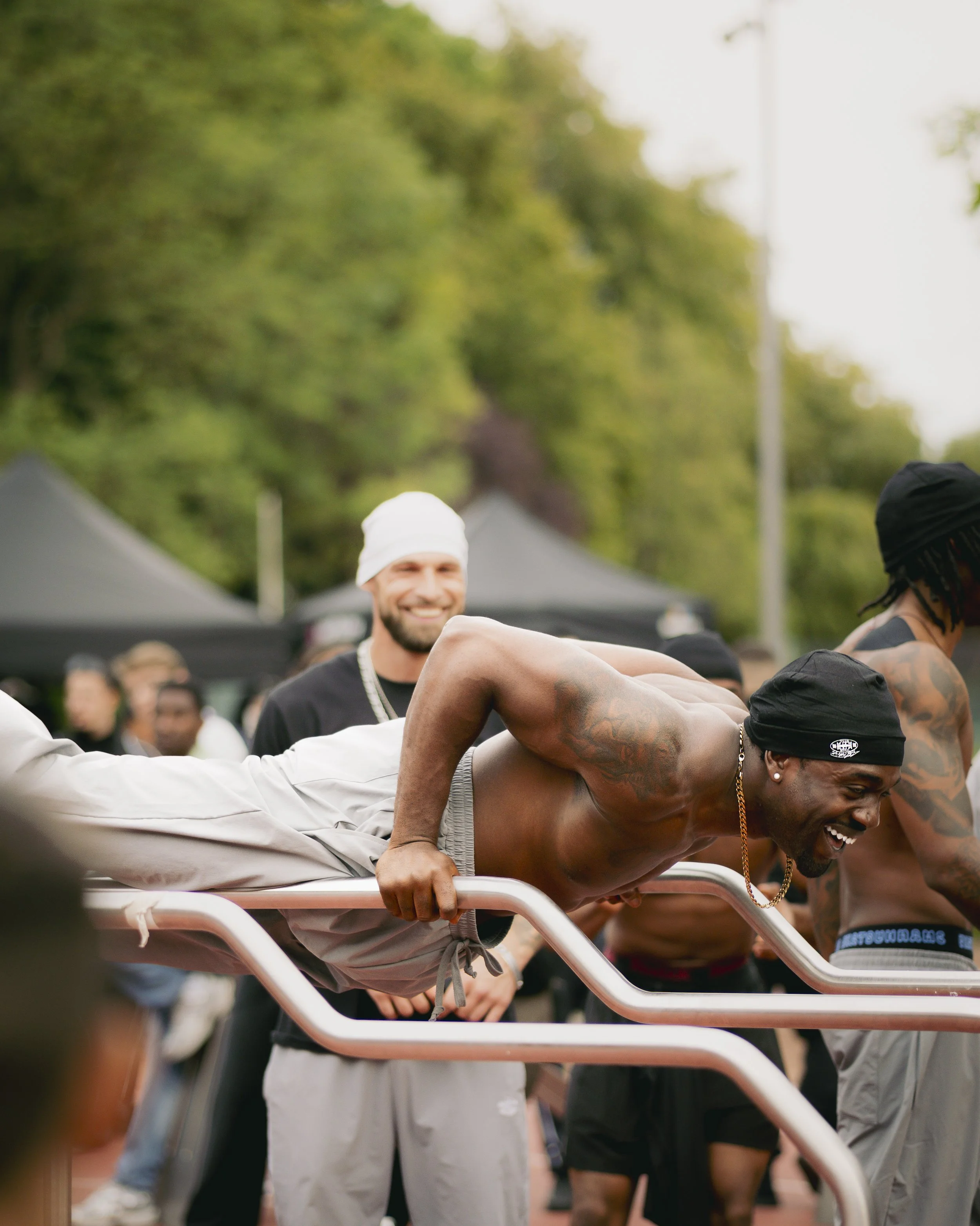 Mehrere Menschen, einschließlich eines schwarzen Mannes, der in einem Calisthenics Park steht, lachen und posieren. Im Hintergrund sind Bäume und Menschen sichtbar.