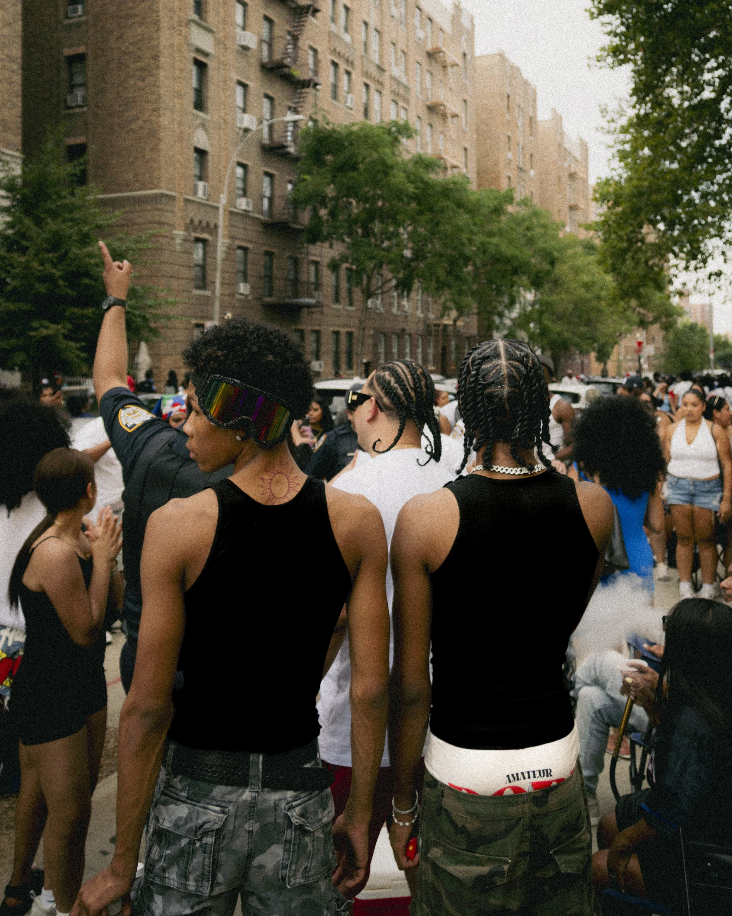 Menschen bei der Dominikanischen Parade in der Bronx New York in einer urbanen Umgebung, mit grauen Hochhäusern und viel Grün im Hintergrund, einige tragen Sonnenbrillen, auffällig ist ein Hand-Emoji auf einem T-Shirt oder Flaggen.