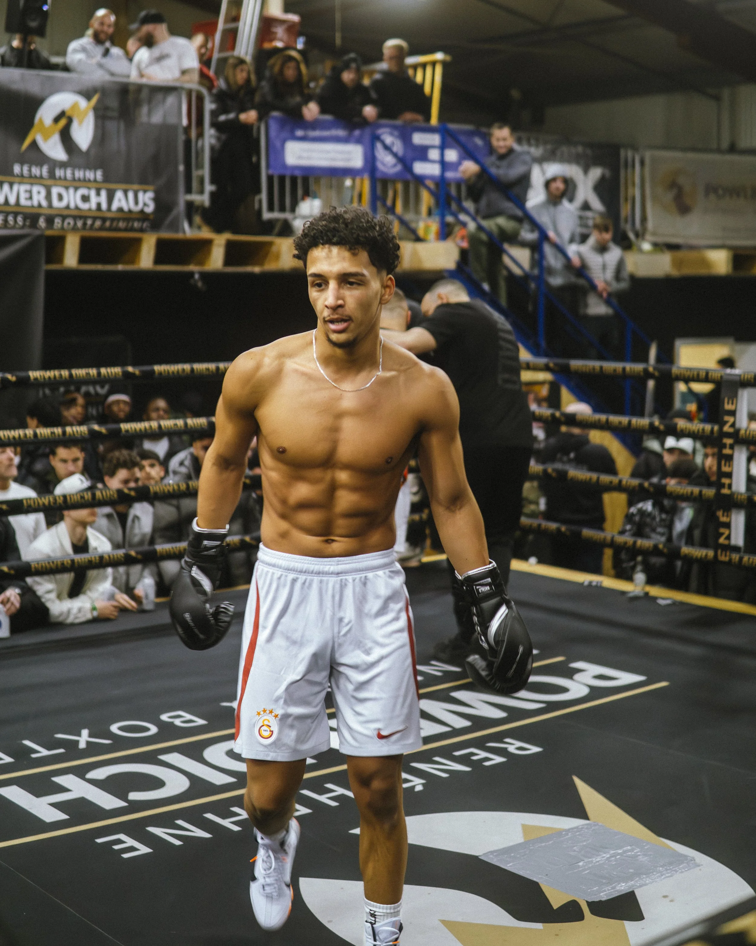 Ein Boxer läuft im Ring; im Hintergrund sitzen Menschen und beobachten das Match, mit Bannern und Absperrgitter im Boxring.