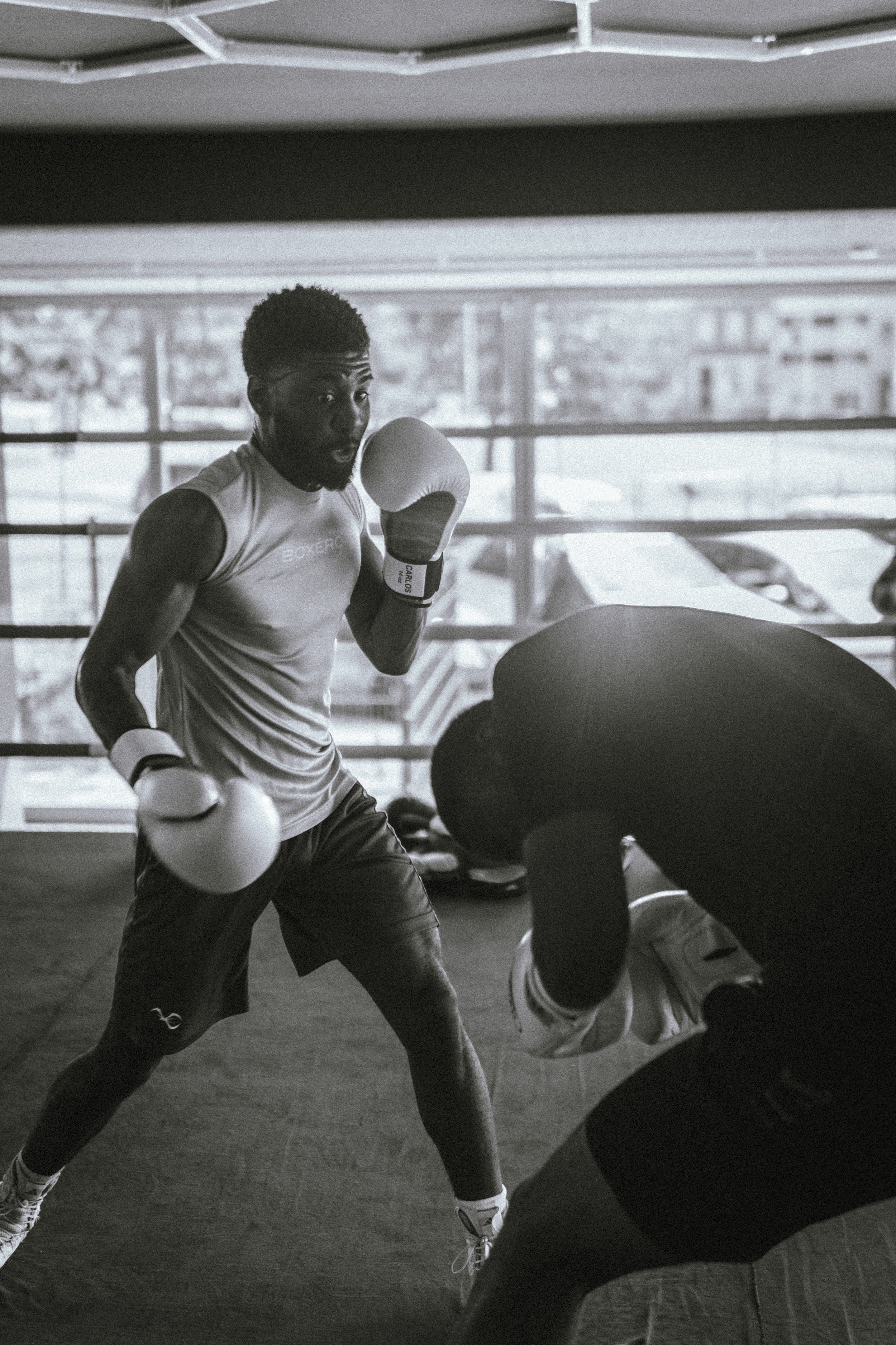 Schwarz-weiß-Foto von einem männlichen Boxer im Training im Fitnessstudio, das er in einer Boxe rkampfform hält, mit einem Boxsack vor ihm und einem großen Fenster im Hintergrund.