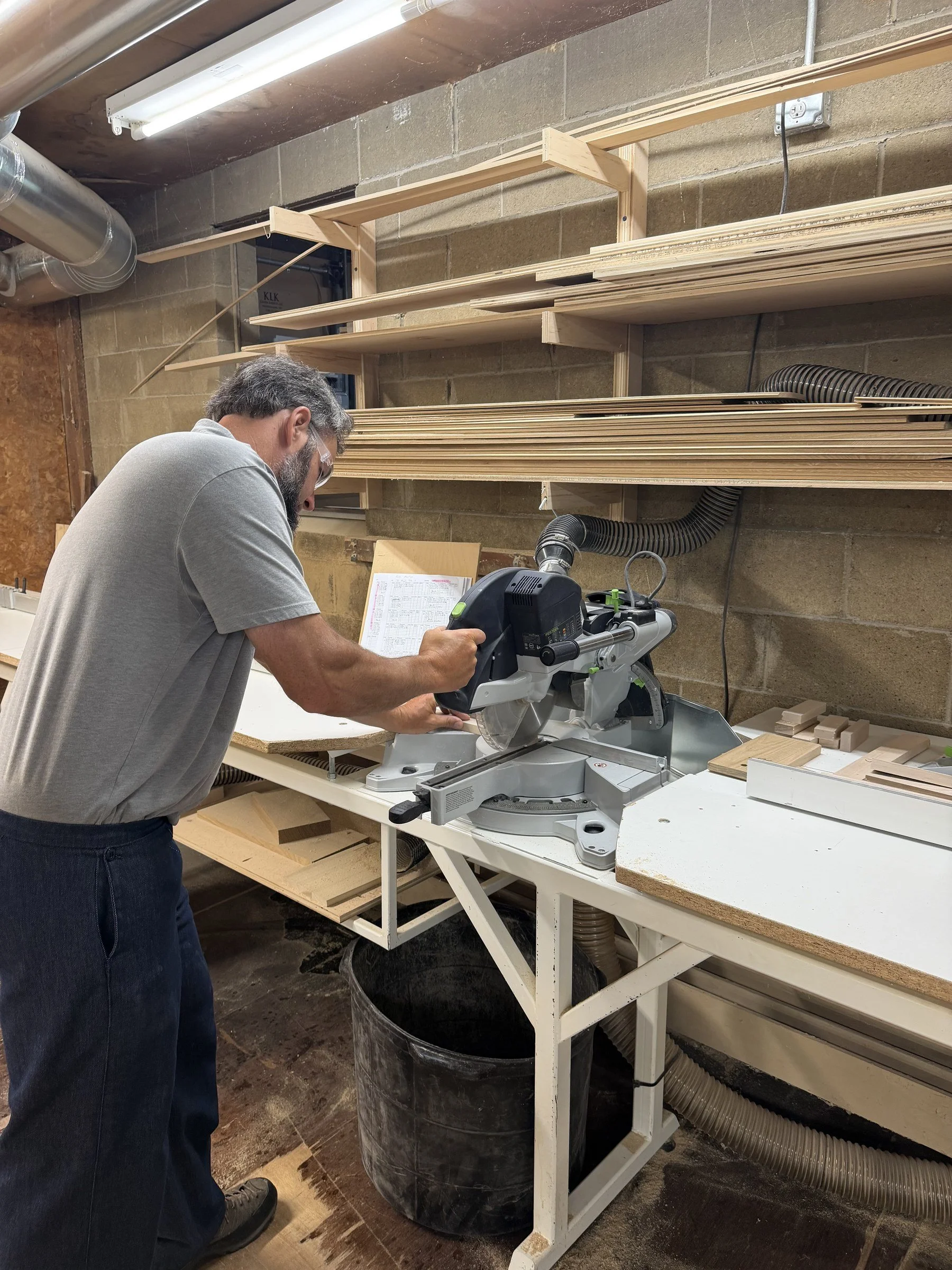 Amish craftsman cutting cabinet stock on a mitre saw, with hardwood lumber racked behind him