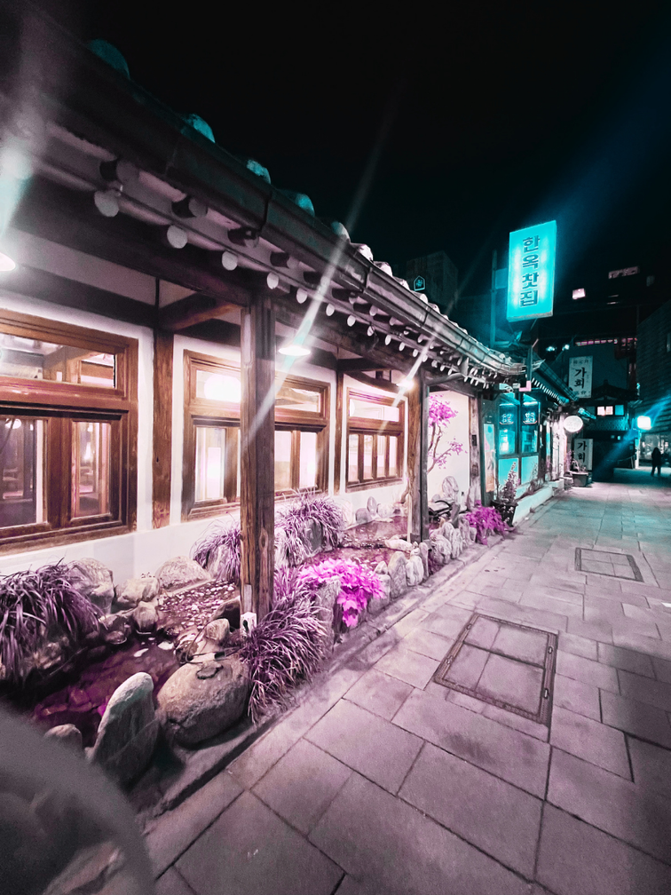 Vista nocturna de un callejón en una zona urbana con un edificio con ventanas de madera, plantas decorativas y letreros iluminados en un idioma asiático