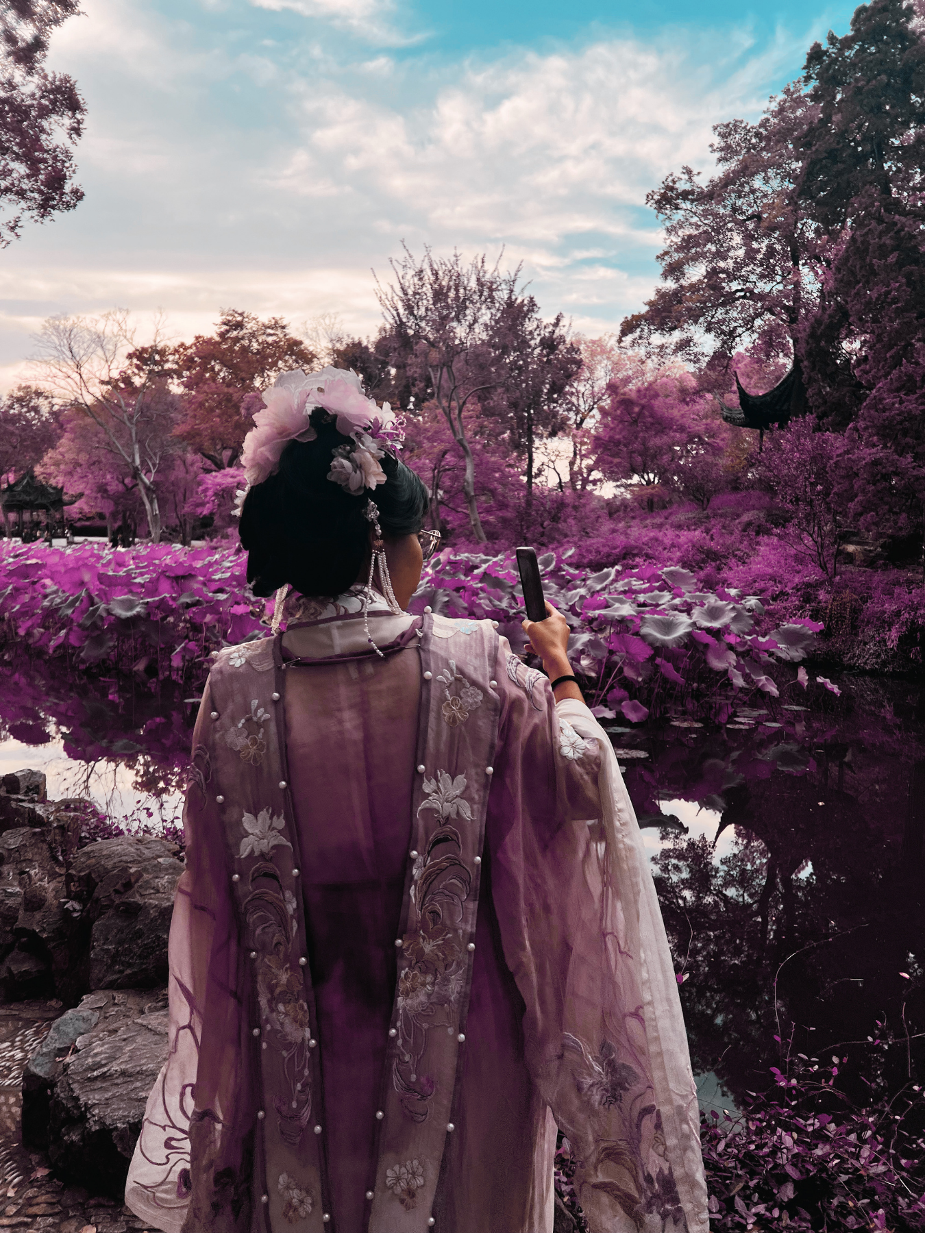 Persona con ropa tradicional japonesa, con un peinado adornado con flores, tomando una selfie en un entorno natural con árboles con flores rosadas y un lago