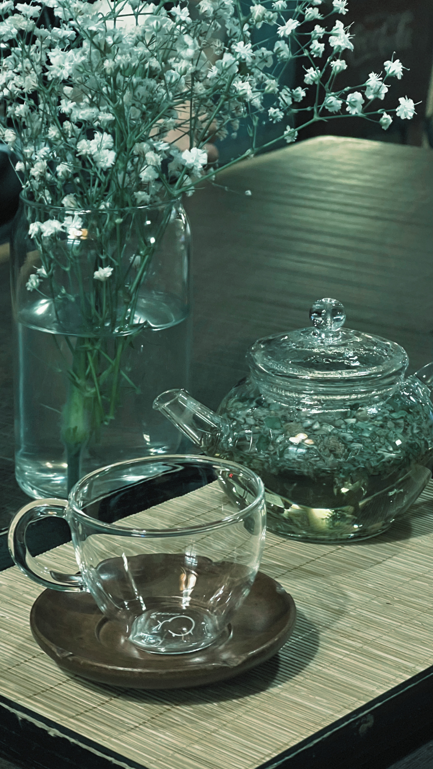 Ramo de flores blancas en un florero, tetera de vidrio con tapa, taza de vidrio con plato, todo sobre una esterilla de madera en una mesa de madera.