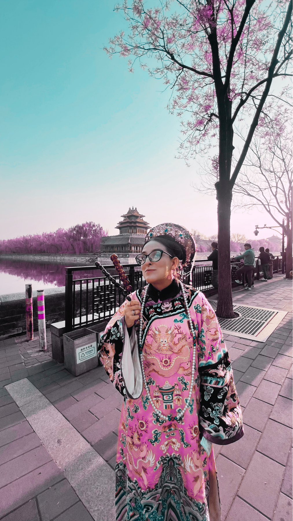 Persona vestida con un kimono tradicional, de pie junto a un río con árboles en flor rosa y un edificio antiguo en el fondo, en un parque urbano en Japón.