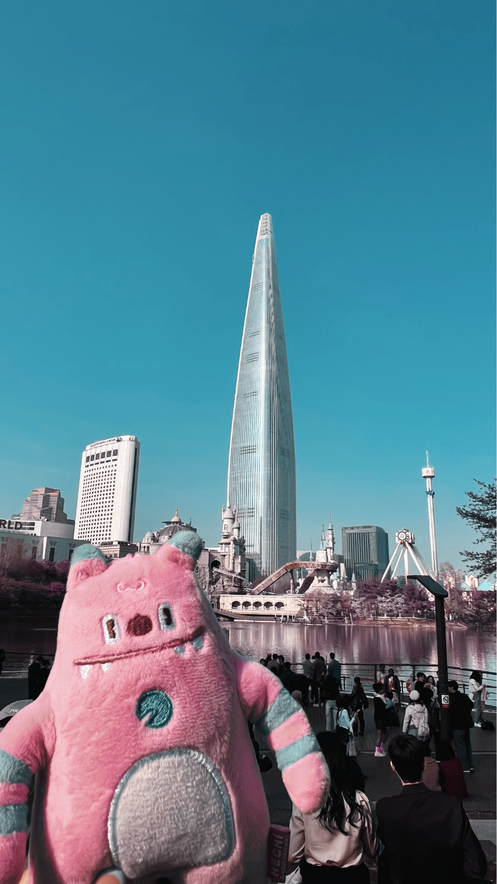La imagen muestra un edificio alto y moderno en un cielo despejado, con una fuente y un parque en primer plano. En el primer plano hay un peluche rosa con detalles en gris y azul, con forma de cerdito.