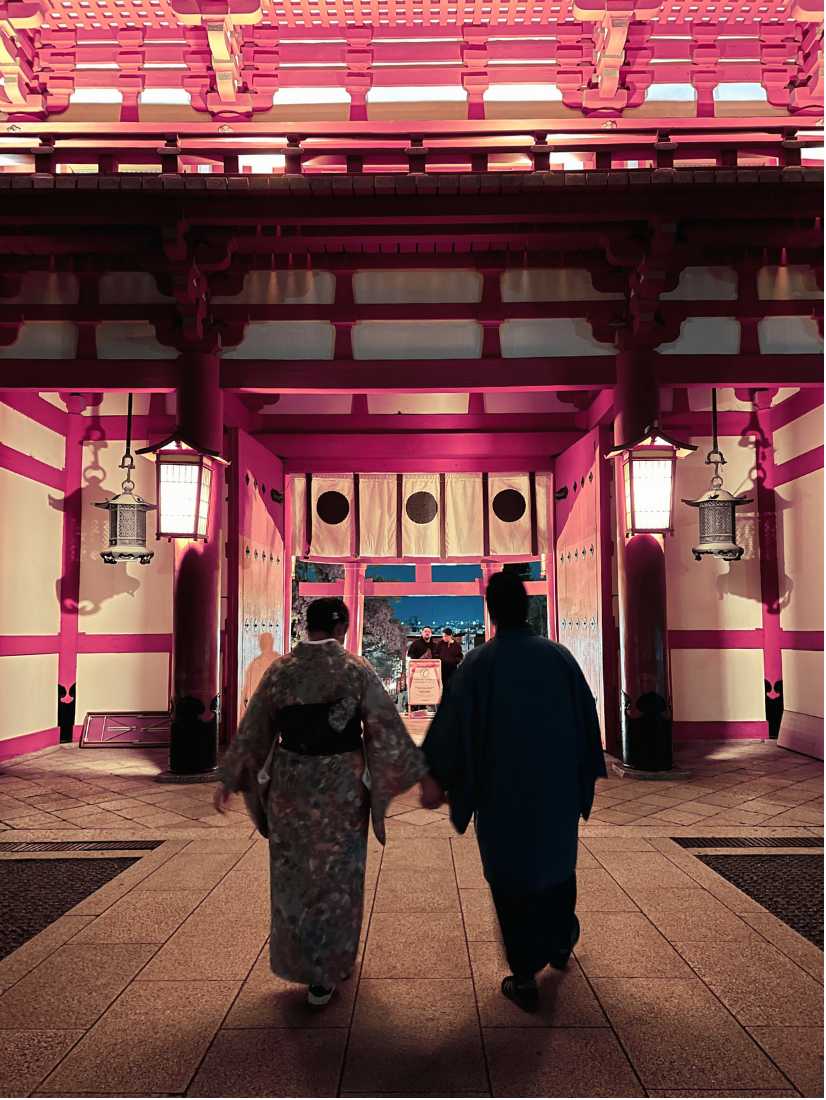 Dos personas con kimono caminando hacia la entrada de un edificio tradicional japonés con estructura de madera pintada de rosa y lámparas en los lados, durante la noche.