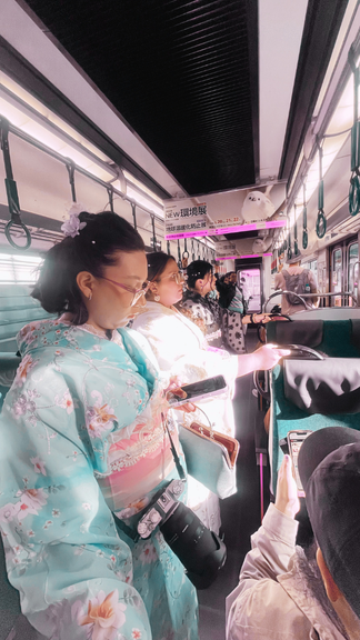 Grupo de personas en un autobús, algunas usando mascarillas y vestidas con kimonos tradicionales japoneses, con posaderas en las ventanas mientras miran sus teléfonos.