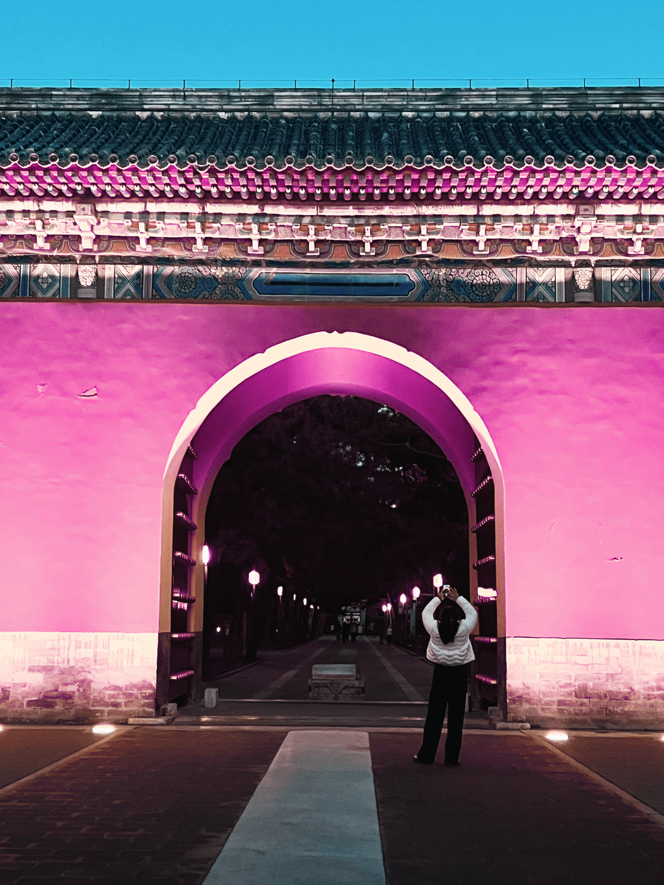 Puerta de estilo tradicional con arco en color rosa y detalles decorativos en la parte superior, iluminada por farolas. Persona tomando una foto desde el arco, en un ambiente nocturno.