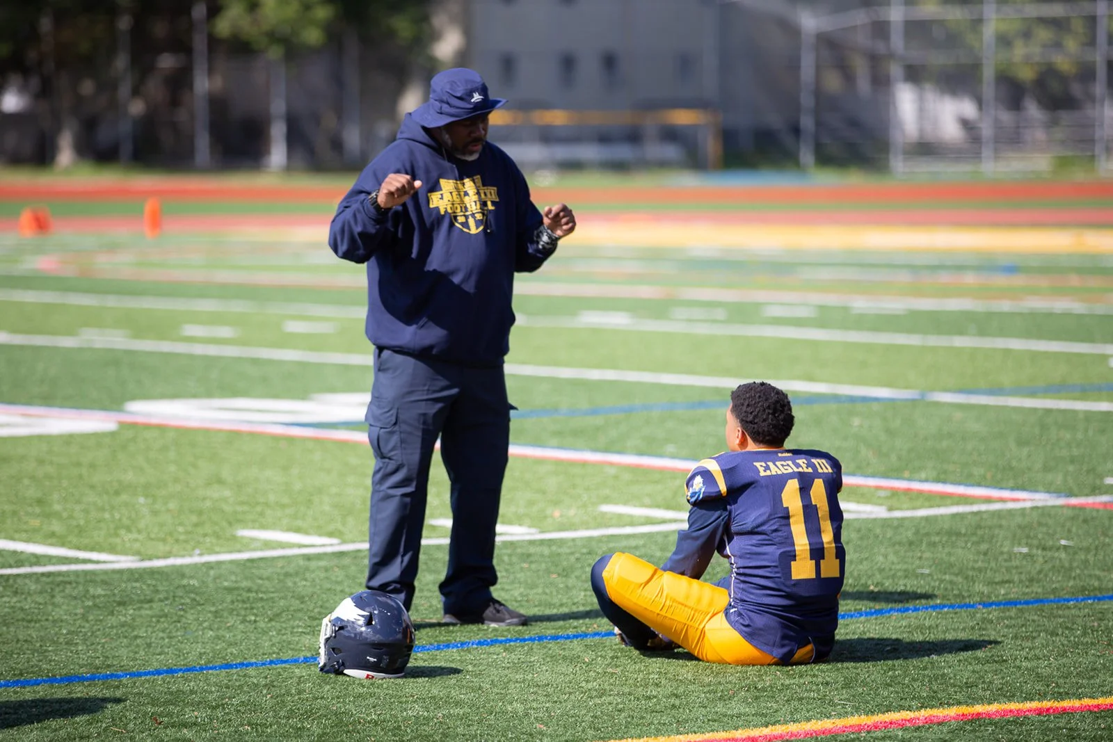 A football coach is talking to a player who is sitting on a football field. The player is wearing a blue and yellow uniform with the number 11 and the name Eagle. The coach is wearing a navy hoodie and a hat, and there is a football helmet on the gro