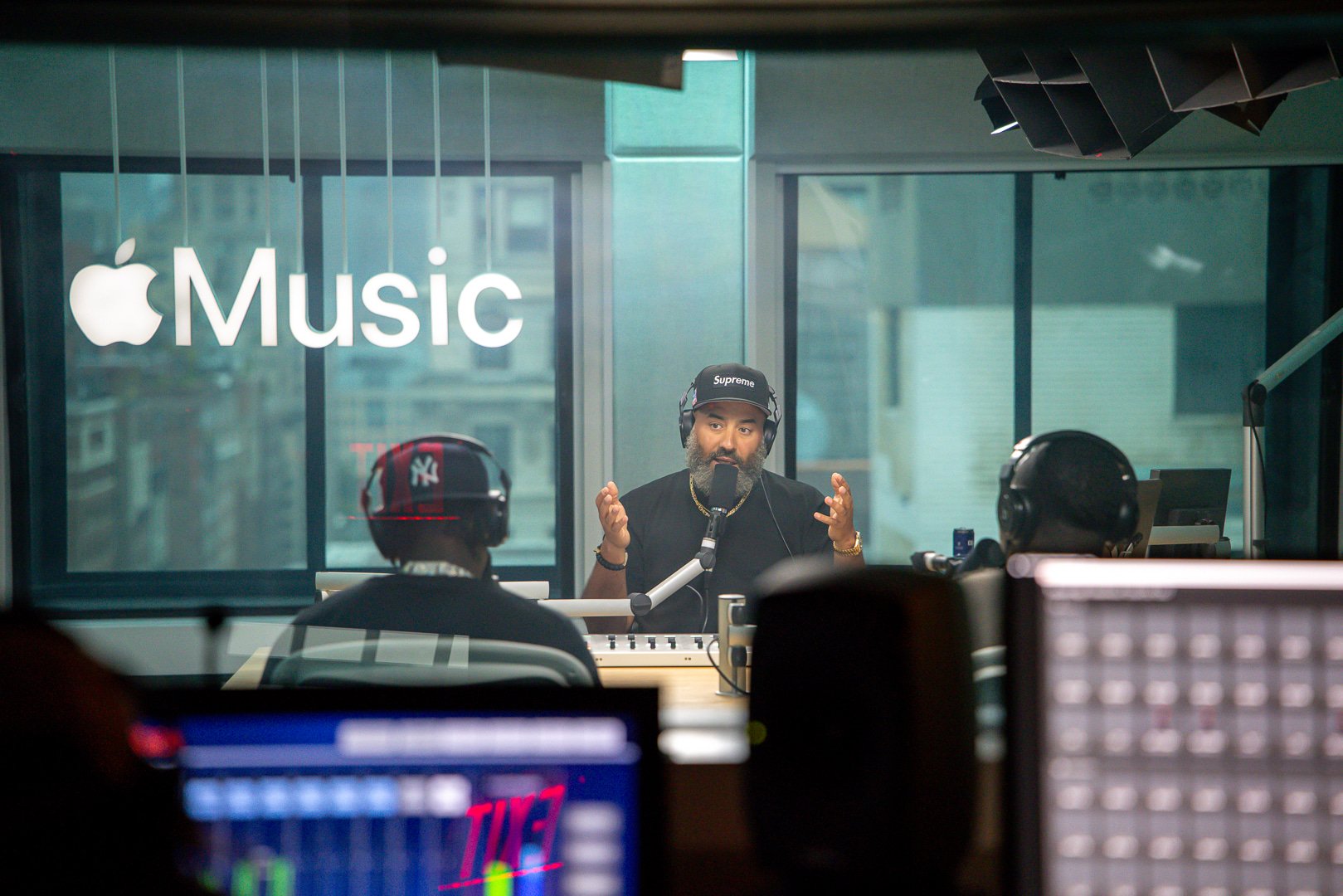 Recording studio with 'Apple Music' sign on window; man with headphones and microphone speaking, three people in front wearing headphones, equipment and screens visible.