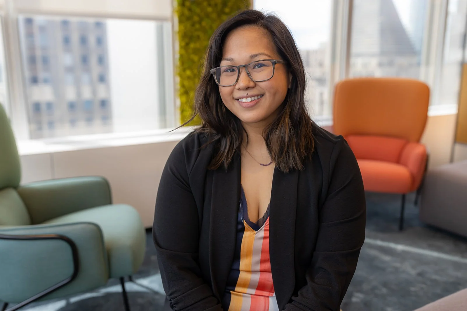 A smiling woman with glasses and shoulder-length dark hair, wearing a black blazer and a multicolored striped top, sitting in a modern office space with large windows and colorful chairs.