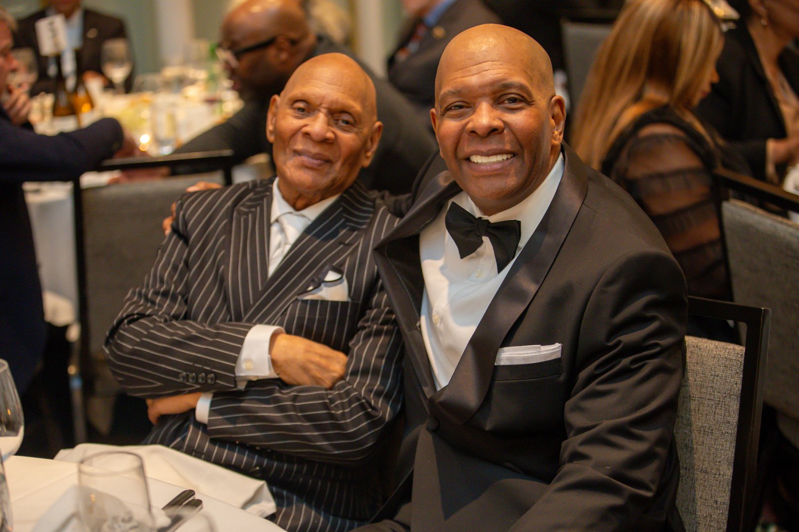 Two men dressed in tuxedos sitting at a formal event, smiling at the camera. The man on the right wears a black bow tie, and the man on the left wears a pinstripe tuxedo.