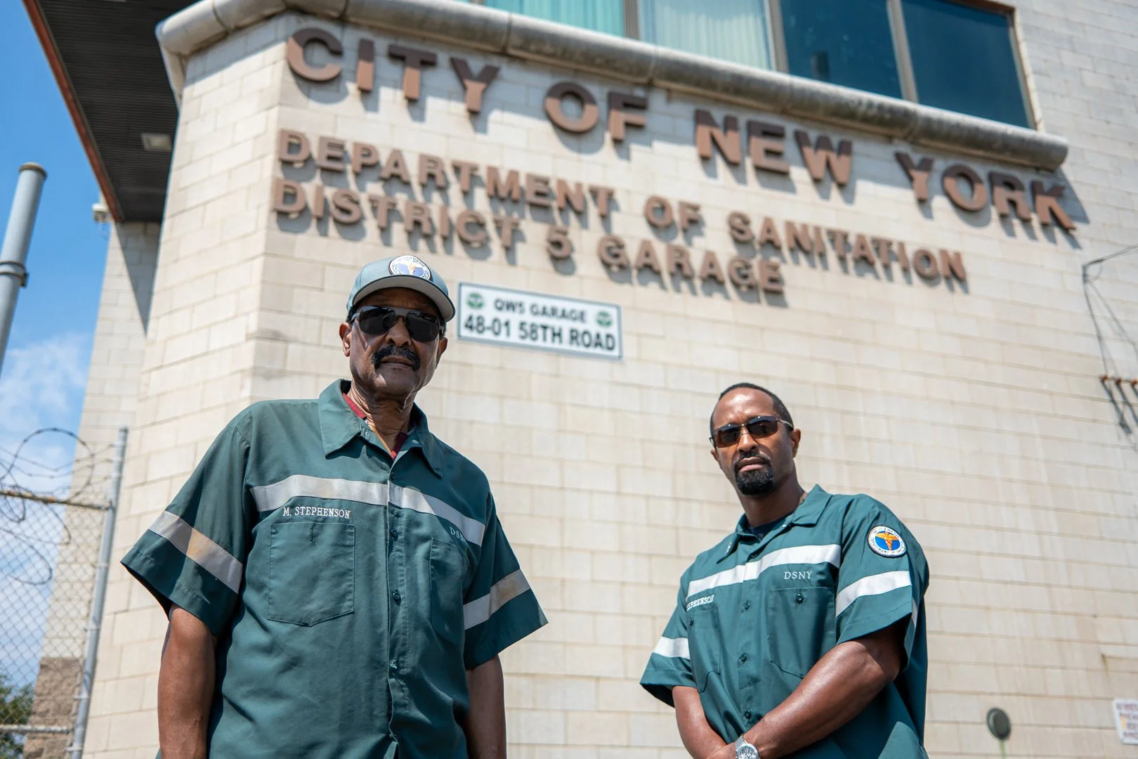 Two uniformed men standing in front of a large building with the sign 'City of New York Department of Sanitation District 5 Garage' and another sign reading 'OWS Garage 48-01 58th Road.' Both men are wearing dark sunglasses, one with a moustache and 